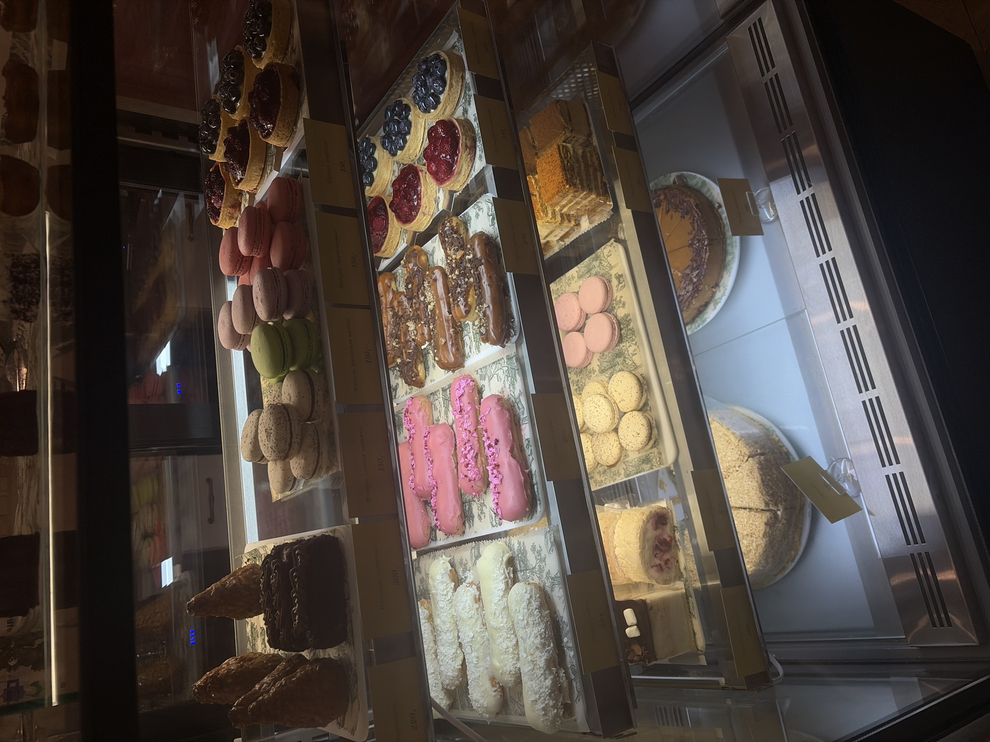The image depicts the interior of a dessert display case, likely within a bakery or cafe in Krasnoyarsk, Russia. No individuals are present.
The display case is multi-tiered, constructed with glass panels, and illuminated internally, creating reflections. The base of the case features horizontal metal grilles.
**Second Shelf from Top:**
* **Left:** Contains four light brown, conical pastries and one rectangular dark chocolate cake with textured frosting on a white tray with a green patterned border.
* **Right:** Displays stacks of various colored macarons: light green, light pink, and off-white. A yellow price tag below indicates "Макарон" (Macaron) and "150 руб." (rubles). Another tag shows "220 руб."
**Third Shelf from Top:**
* **Left:** Features eight elongated pastries (eclairs), uniformly coated in white icing; some are topped with shredded coconut. These are on a white tray with a green patterned border.
* **Right:** Shows eight elongated pastries (eclairs), uniformly coated in pink icing; some are decorated with pink sprinkles. These are on a white tray with a green patterned border. Price tags beneath these sections indicate "200 руб." and "190 руб."
**Fourth Shelf from Top:**
* **Left:** Holds several dark-glazed elongated pastries (eclairs) topped with crushed nuts or chocolate pieces, arranged on a white tray with a green patterned border.
* **Right:** Presents multiple small, circular tarts with pastry crusts. Some are filled with red berries (e.g., raspberries, cranberries), while others are filled with dark blue berries (e.g., blueberries, blackcurrants). These are on a white tray with a green patterned border. A price tag below indicates "150 руб."
**Fifth Shelf from Top:**
* **Left:** Contains several light pink macarons on a white tray with a green patterned border.
* **Middle:** Features at least two multi-layered rectangular cakes, possibly honey cake (Medovik), and a white roll cake with a red fruit filling visible. A small white square dessert topped with a marshmallow is partially visible.
* **Right:** Displays several off-white macarons.
**Bottom Shelf:**
* A large, round, sliced cake with a light-colored, crumbly topping is visible on a white plate. Another round, sliced cake with a darker topping is also visible on a white plate.
The background, visible through reflections in the glass, suggests a retail environment with other display units or shelving. The overall scene is well-lit for product display within the case, contrasting with a slightly dimmer surrounding environment.