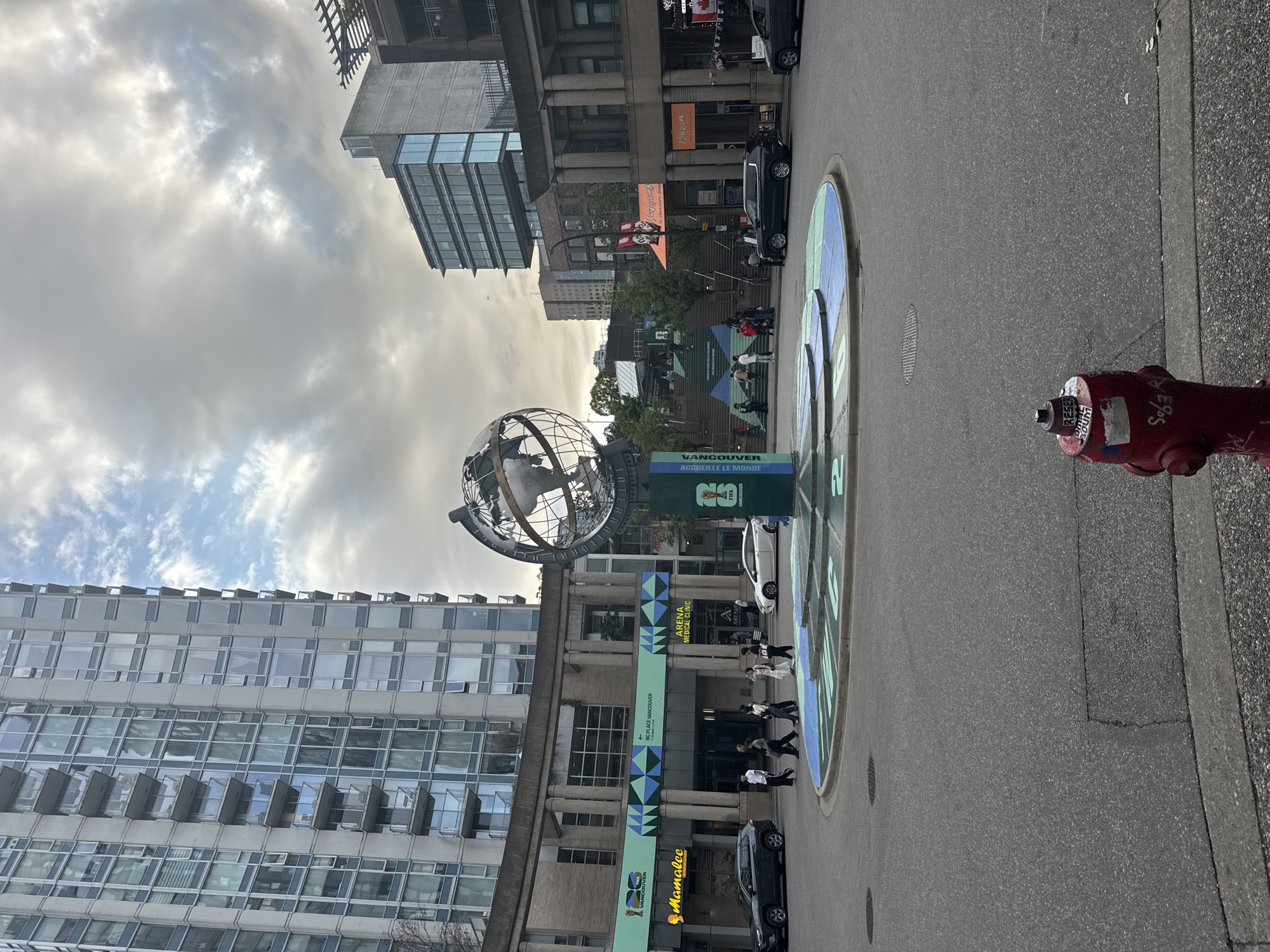 This image depicts a wide-angle view of a city street in Vancouver, Canada, dominated by a large, modern globe sculpture at an intersection. The sculpture is the focal point, with its metallic framework clearly showing continents and oceans. It stands on a circular, multi-colored base that is painted onto the asphalt.
To the left of the sculpture is a tall, modern building with a glass facade and balconies. Below this building, at street level, is a row of shops and establishments. Prominently visible are signs for "Mamalee" and "ARENA MEDICAL CLINIC". A large green banner with blue diamond patterns and the text "BC PLACE VANCOUVER" and "HOME OF THE LIONS" is also displayed. Several people are walking on the sidewalk in front of these establishments, and cars are parked or driving on the street.
To the right of the globe sculpture, the scene continues with more buildings and shops. An orange awning is visible, along with more signage, including a Canadian flag. A set of outdoor stairs with colorful geometric patterns leads upwards, with people visible on them. Further in the background, more contemporary buildings can be seen, including one with a distinctive glass facade.
The sky above is overcast with scattered clouds, suggesting a potentially cool or temperate day. The overall impression is that of a vibrant urban environment, likely a public square or a busy street corner in Vancouver, Canada, possibly near BC Place Stadium given the signage. A bright red fire hydrant is positioned in the lower right foreground of the image.