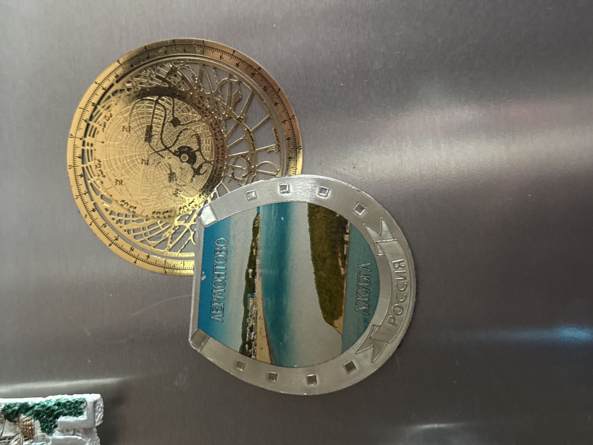 The image displays two magnets on a metallic surface, possibly a refrigerator door. The magnet on the left is a circular, golden astrolabe with intricate celestial patterns and markings. The magnet on the right is shaped like a horseshoe and features a scenic view of a beach with turquoise water, a sandy shore, and lush green hills. Text is visible on the horseshoe magnet. On the left side, in blue Cyrillic, it reads "АЕРМОНТОВО" (Aermontovo). On the right side, also in blue Cyrillic, it reads "ДЖУБГА РОССИЯ" (Dzhubga Russia). The scene is indoors, with the metallic surface suggesting a kitchen setting. The lighting is neutral, and there are no clear indicators of the time of day or weather conditions. The overall impression is of decorative souvenirs. There is also a small, partially visible magnet in the bottom left corner, appearing to be a miniature white building with green detailing.