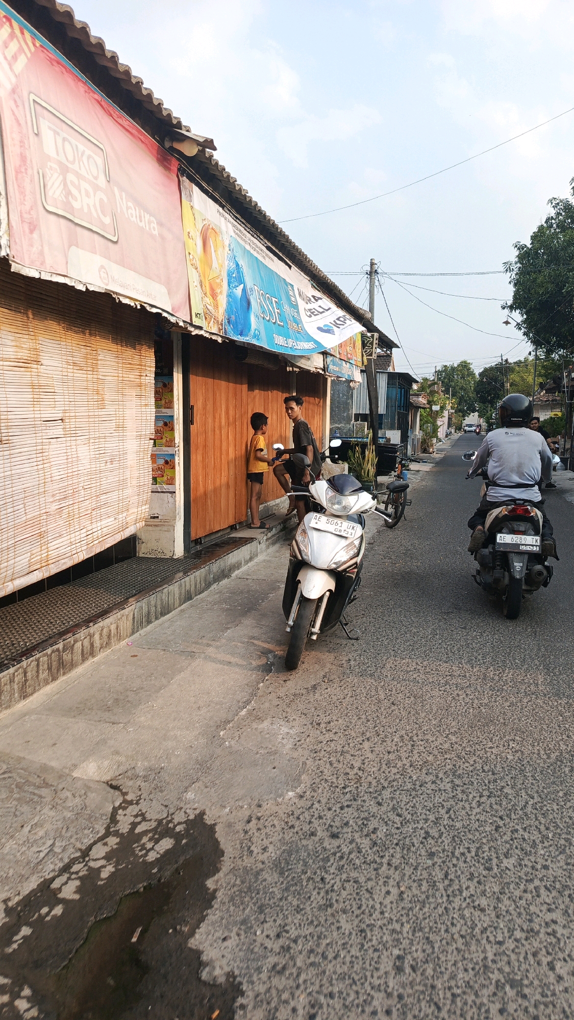 In the city of Mangunsuman, Indonesia, a street scene unfolds with a row of shops lining the left side of a paved road. A banner advertising "Toko SRC Naura" with the tagline "Melayani Pesan Antar" stretches across the top of one shop. The shopfront features a wooden door and a bamboo blind covering a window.
Two young men are engaged in conversation near the entrance of the shop. A white motorcycle is parked on the sidewalk in front of them. Further down the street, two other motorcycles are visible. One is parked on the sidewalk, and another is being ridden by a person wearing a helmet and a light-colored shirt, moving away from the camera. The road is asphalt and appears wet in patches. Trees and some buildings can be seen in the background, suggesting a semi-urban or suburban environment. The sky is clear and blue, indicating daytime.