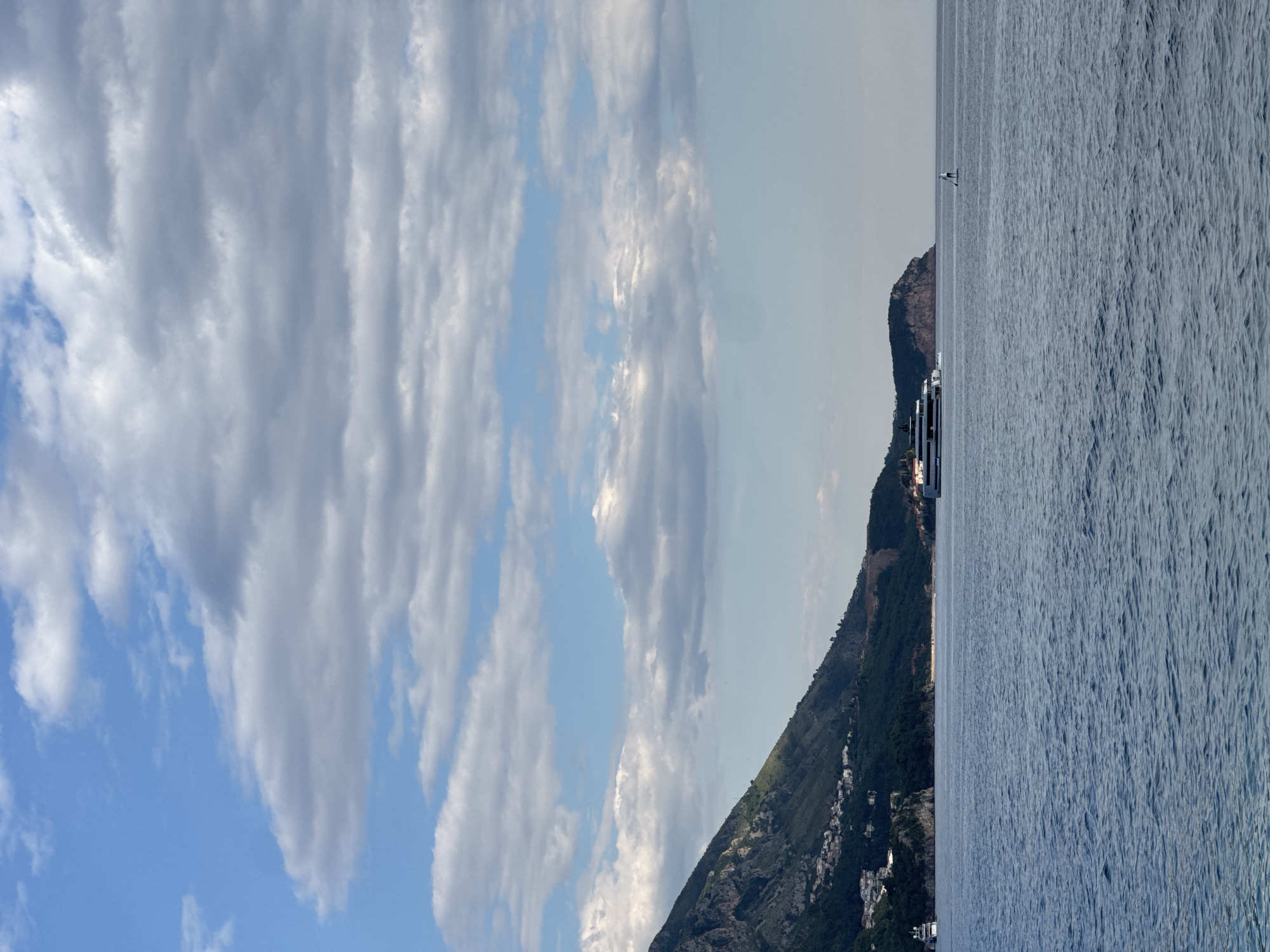 The image depicts a coastal scene in Rafailovići, Montenegro. A mountain with dense vegetation and scattered buildings slopes down to the sea. A large cruise ship is anchored near the coast, and a person is visible in the water, possibly swimming or paddleboarding. The sky is partly cloudy with patches of blue. The sea is calm with slight ripples on the surface.