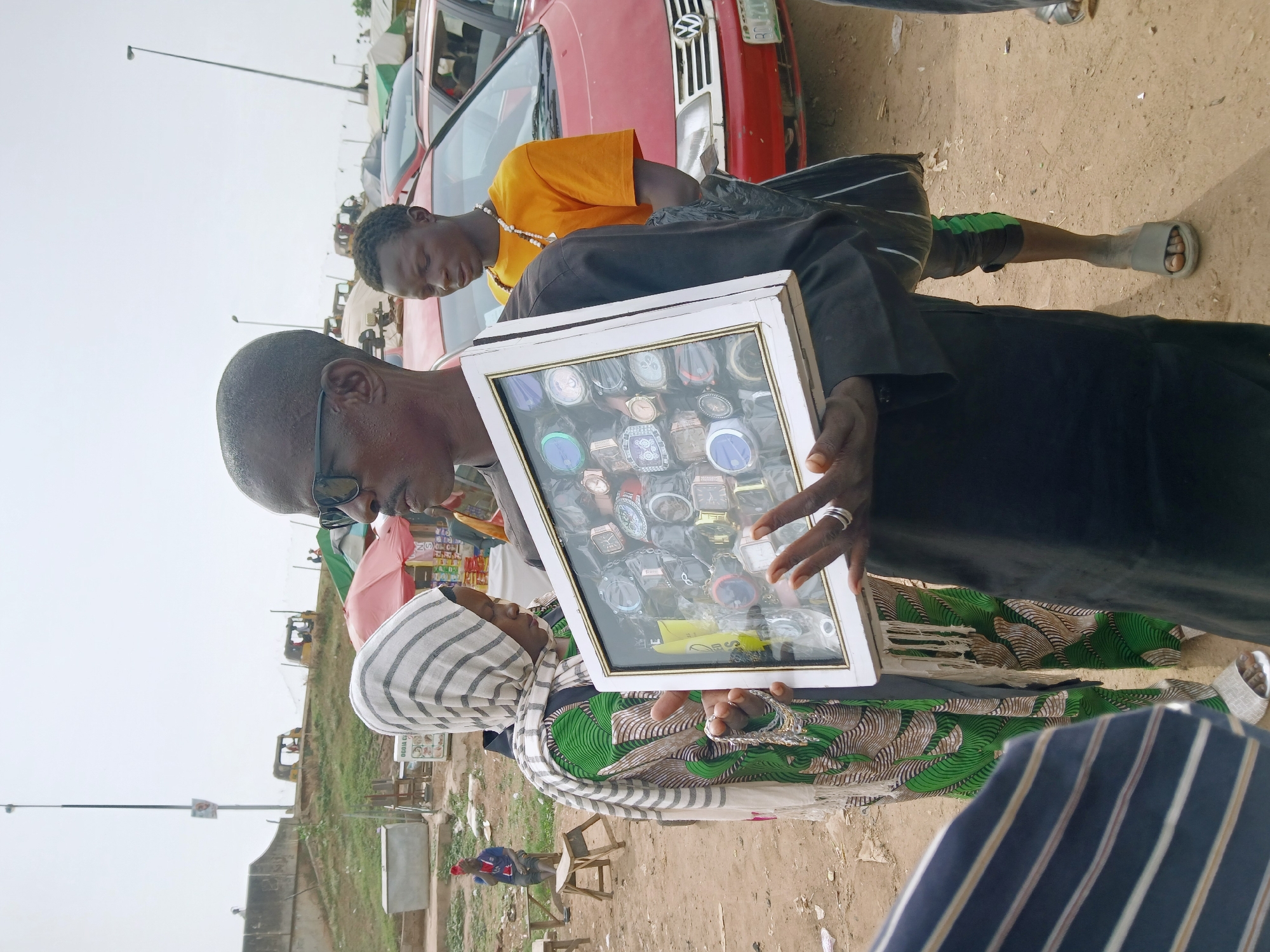 In an urban setting, a man wearing glasses and a black robe is displaying a case of watches to a woman with a white and black striped headscarf and a green and brown patterned dress. Another man in an orange shirt stands behind them. A red car is visible in the background. The ground appears to be sandy. The location is Unknown, Unknown.