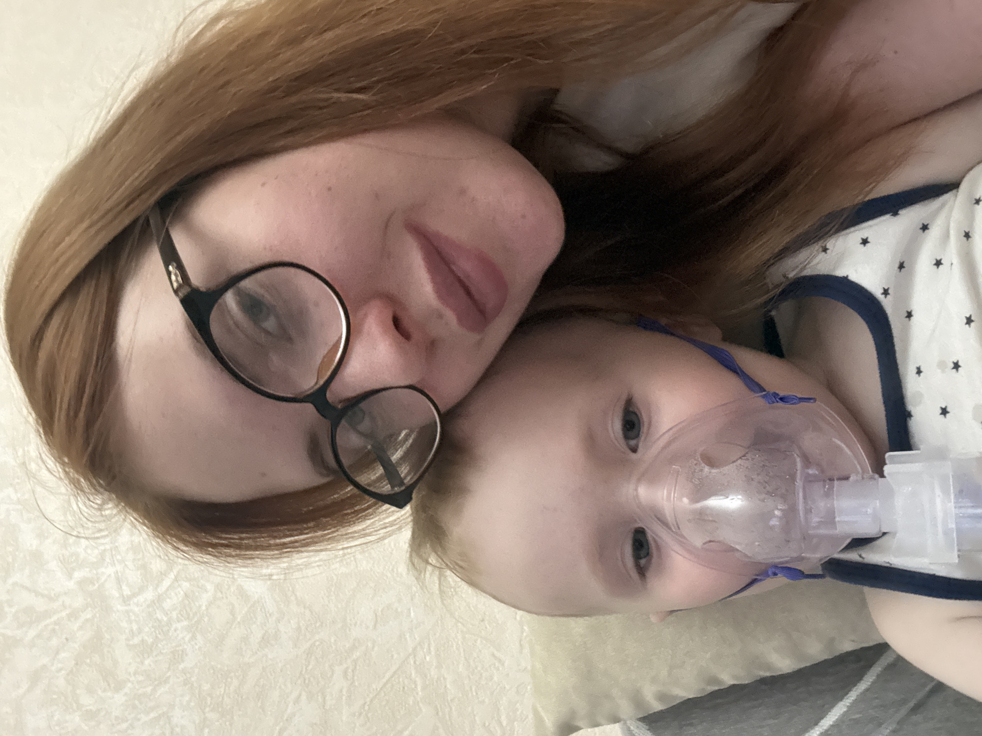 A photograph taken indoors features an adult with long red hair wearing black framed glasses and a young child with an oxygen mask. The adult is lying on a light-colored, textured surface, possibly a bed or couch, with their head tilted slightly towards the camera. The child is positioned similarly next to the adult, with the oxygen mask covering their nose and mouth. The child is wearing a white garment with navy blue trim and a pattern of stars. The setting appears to be a domestic environment, likely a home. The location is Chaikovsky, Russia.