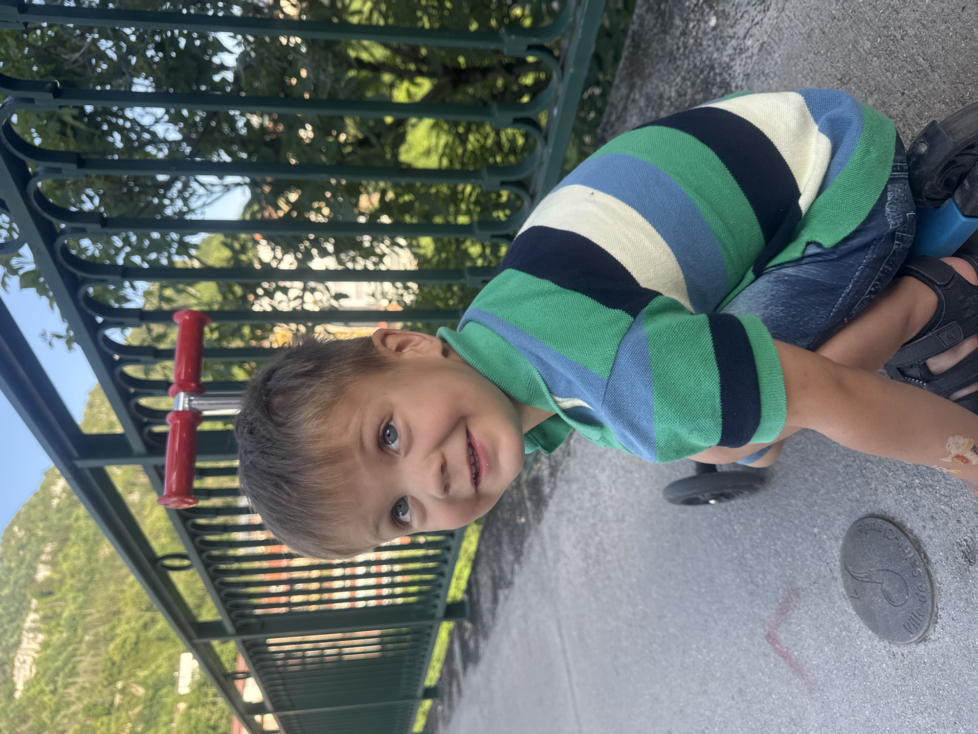 A young boy with short brown hair and blue eyes is pictured from the chest up, looking up and to his left. He is wearing a green, blue, and white striped polo shirt and denim shorts. He is sitting on a blue and black scooter. The background features a green metal fence and a hill with trees and buildings. A small, oval metal plaque is visible on the ground in the lower left quadrant of the image. The plaque has an inscription that reads "Ville de Saint-Claude" and depicts a musical note symbol. This indicates the location is within the city of Saint-Claude, France. The setting appears to be outdoors, likely a park or a pedestrian walkway.