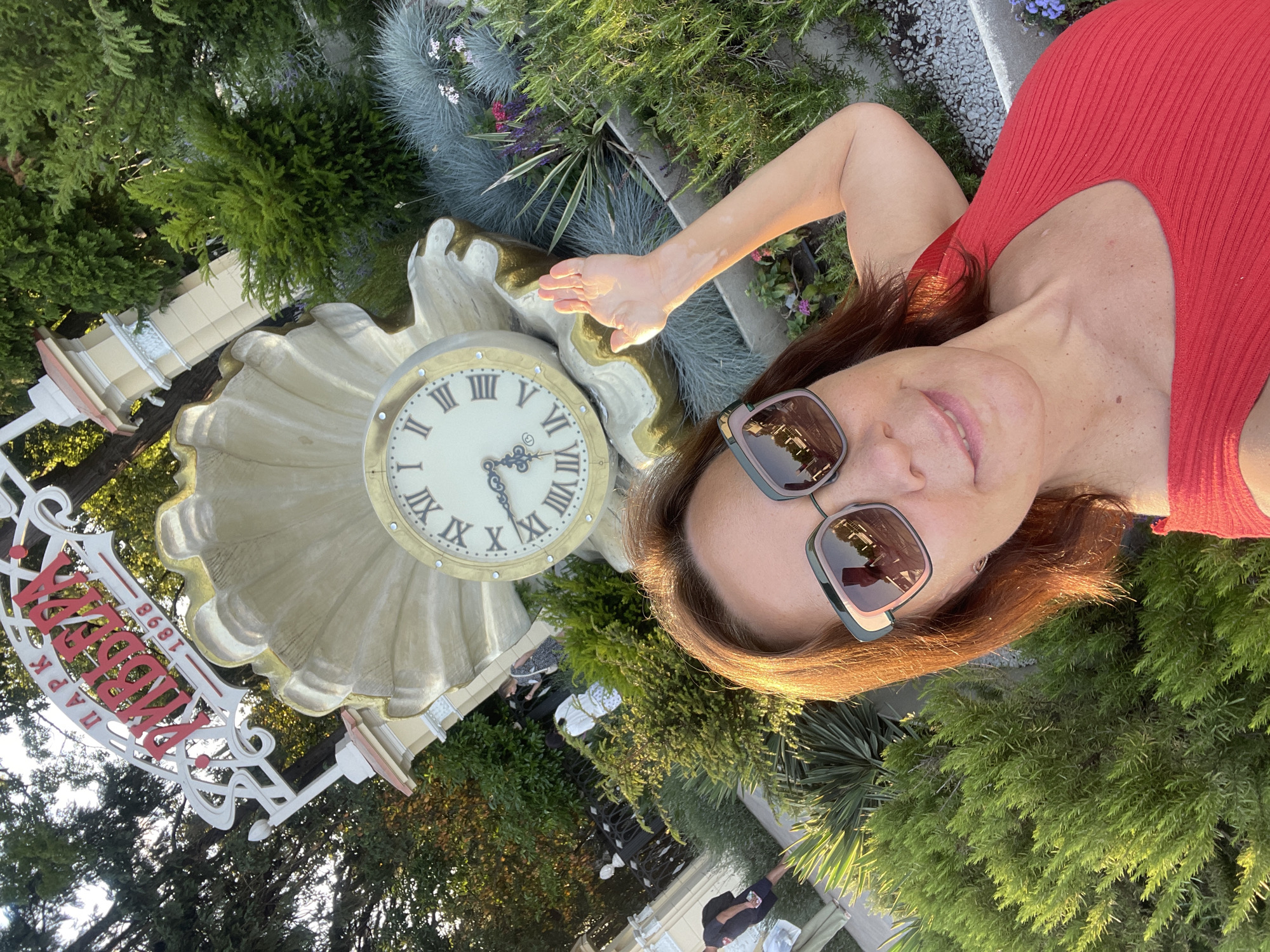A woman with long brown hair and sunglasses is taking a selfie in a park. She is wearing a red top. To her left is a large, ornate clock with Roman numerals. Behind the clock is lush greenery, including various trees and plants. In the background, there is a sign with red lettering and the year "1898". The sun is shining, casting a warm glow on the scene. The image appears to have been taken during the daytime. The woman's expression is relaxed and happy. The overall setting suggests a pleasant outdoor environment, likely a tourist attraction or a well-maintained park. The text on the sign appears to be in Russian, possibly indicating the name of the park and its founding year.
