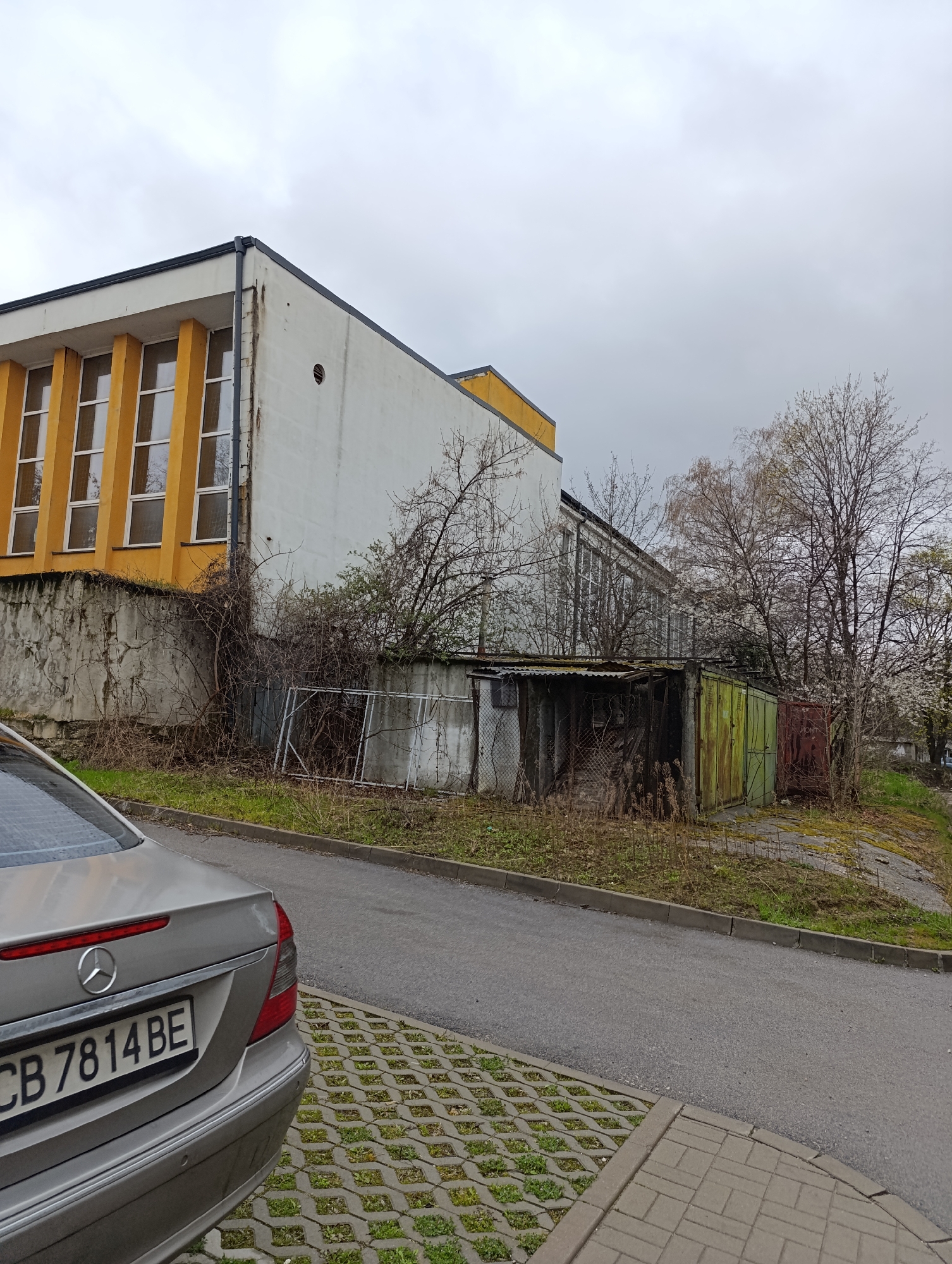 The image depicts a street scene in Sofia, Bulgaria, with a silver Mercedes-Benz car partially visible in the lower left corner. Its license plate reads "CB 7814 BE". The background features a dilapidated, two-story building with a yellow and white facade. Adjacent to the building are a series of deteriorating garages with varied colors including white, green, and red. Overgrowth and bare trees are present between the building and garages. The sky is overcast. The road is paved, and a permeable paving surface with grass growing through it is visible.