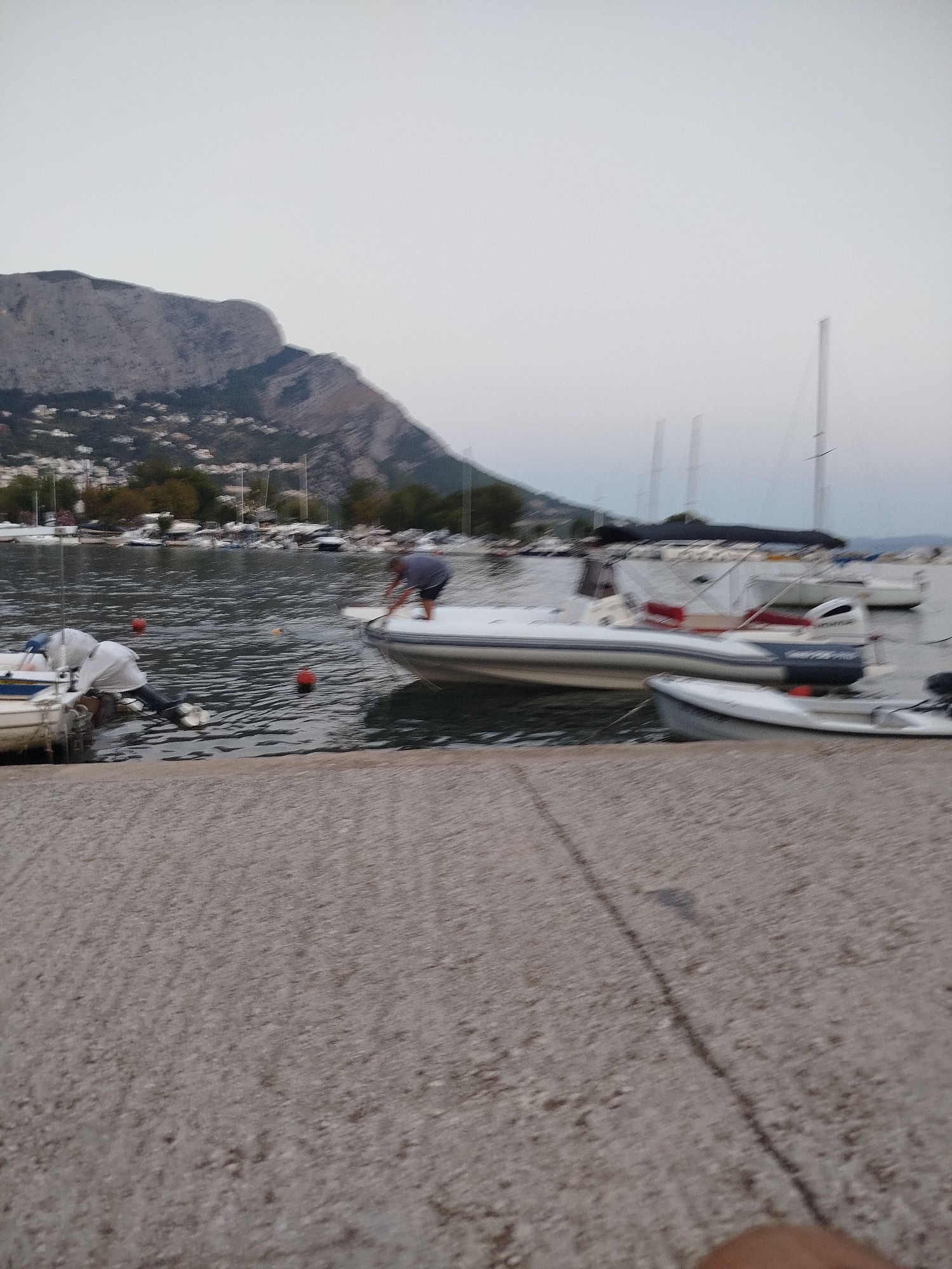 A person is standing on a white inflatable boat with red trim, leaning forward and appearing to adjust something. The boat is in the water next to other boats and a stone embankment. In the background, a mountainous coastline with scattered buildings and trees is visible under a hazy sky. Several sailboats with tall masts are also present in the harbor. The scene is blurry, suggesting motion or a lack of focus. The location is identified as the city of Omiš, Croatia.