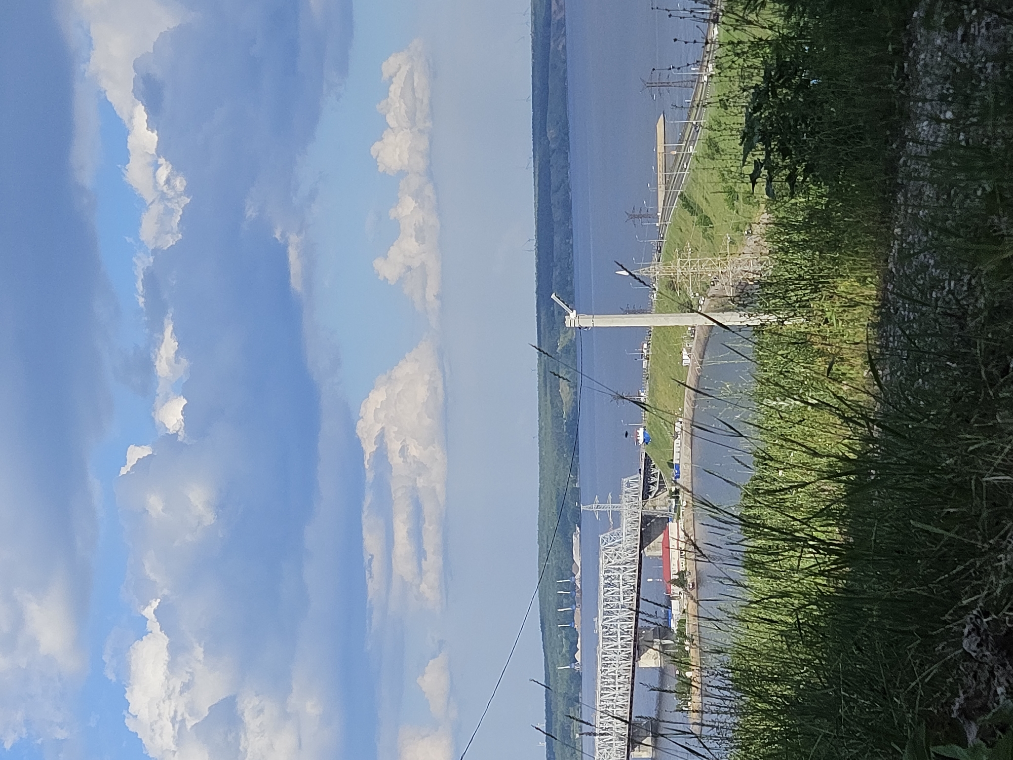 The image captures a wide view of the Volga River and its surroundings in Ulyanovsk, Russia. The scene is divided vertically, with the left side dominated by a bright blue sky filled with fluffy white clouds, suggesting a pleasant daytime. The right side of the image shows the river, a grassy embankment, and parts of an industrial or infrastructure complex.
On the river, a long, flat-bottomed barge is visible, loaded with what appear to be sand or gravel. Further in the distance, a large white bridge with a complex lattice structure spans the river, connecting its banks. Along the embankment, power lines and what seem to be electrical substations or transmission towers are present, indicating industrial activity. The embankment itself is covered in green grass, with some areas appearing more overgrown with taller grasses and plants in the foreground.
The time of day appears to be midday or early afternoon, given the bright sky and sunlight. The weather is fair, with clear skies and abundant sunshine. The location is clearly Ulyanovsk, Russia, indicated by the presence of the Volga River and its characteristic bridge, often referred to as the Imperial Bridge. There are no visible people in the image, nor is there any discernible text. The overall mood of the image is one of natural beauty juxtaposed with significant man-made structures, capturing a slice of life on a major Russian river.
