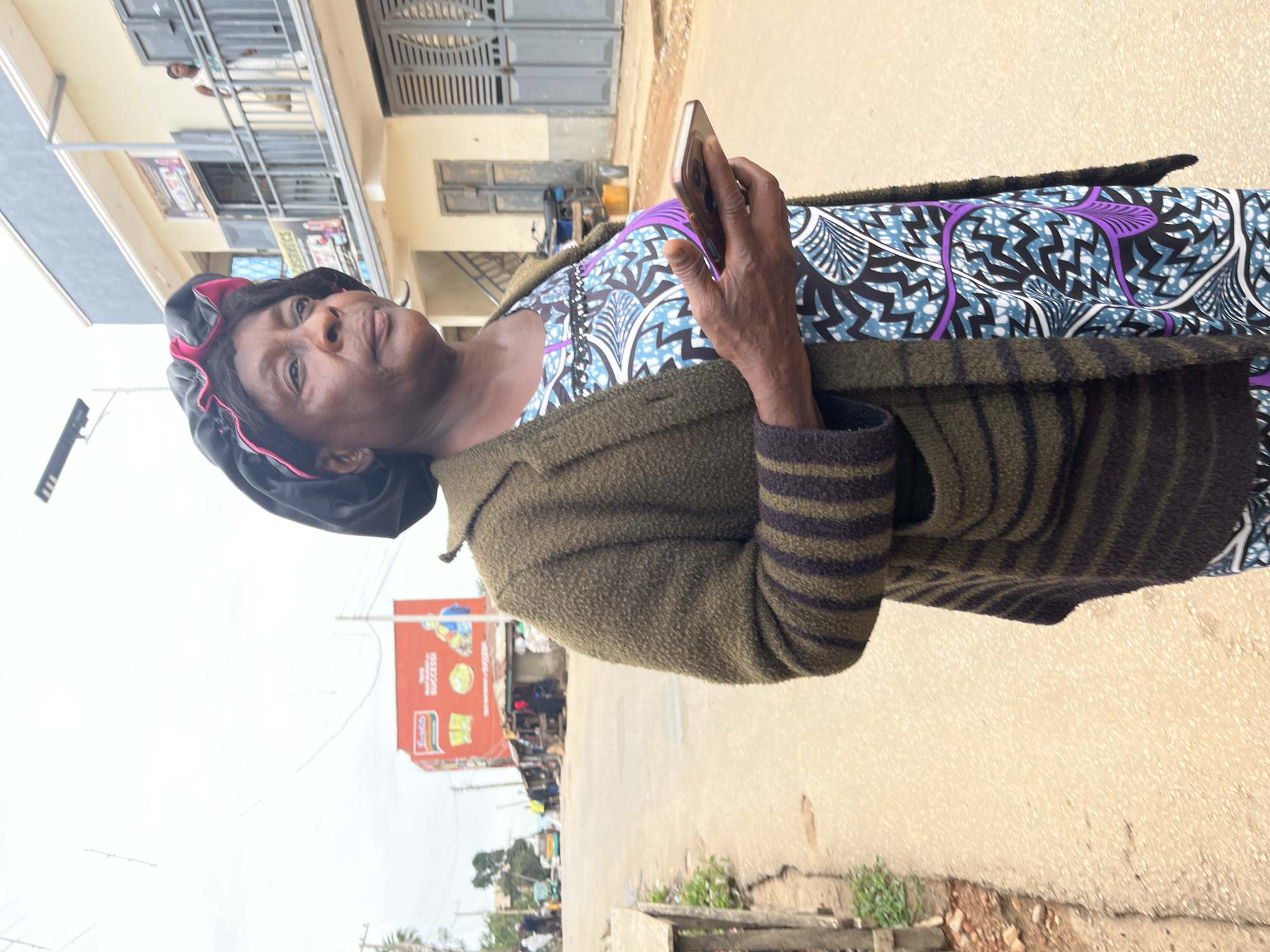 An elderly Black woman, wearing a black satin bonnet with pink trim, a green knitted cardigan with dark stripes, and a blue and purple patterned dress, stands on a paved street in Jos, Nigeria. She holds a smartphone in her right hand. In the background, a two-story building with balconies and closed shutters is visible, along with a large red billboard advertising "Indomie Success." Several people and vehicles are present further down the street. The sky is overcast.