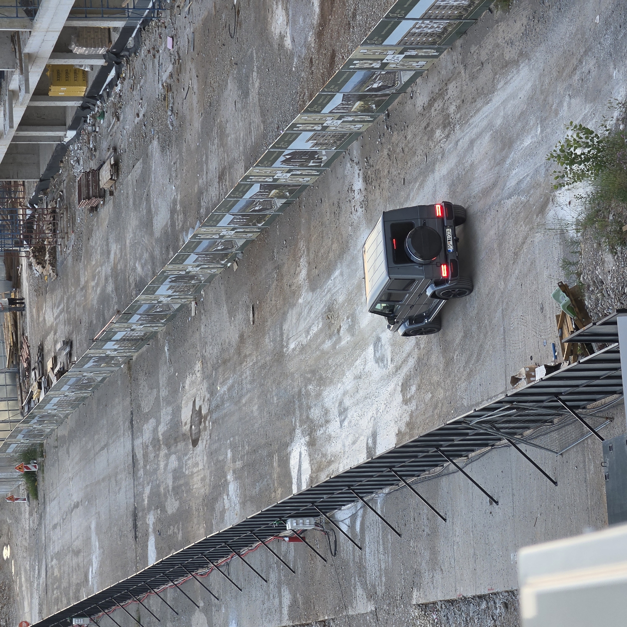 The image shows a construction site in Podgorica, Montenegro. A dark-colored Mercedes-Benz G-Class vehicle with license plate "PG-WAGON" is parked on a dirt road between two concrete walls. A metal fence runs along the top of both walls. Debris and construction materials are visible on the left side of the road. Vegetation is present on the right side. Construction infrastructure is partially visible in the background.