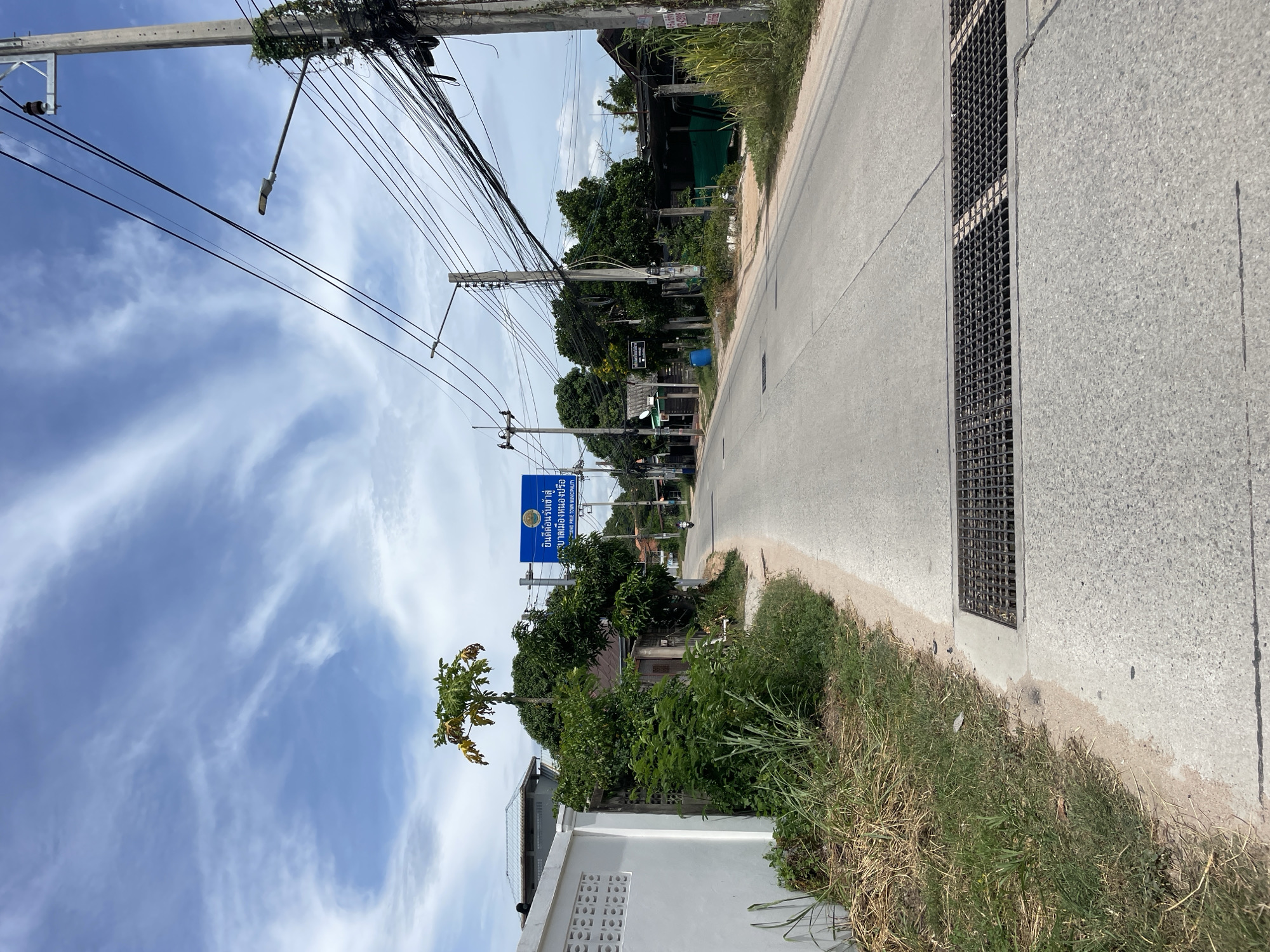 The image shows a street in the city of Muang Pattaya, Thailand, on a cloudy day. The street is paved and appears to be a two-lane road. On the left side of the road, there is a white wall, overgrown with grass and weeds. Further down the road on the left, there are several trees and some residential buildings. On the right side of the road, there are more trees, utility poles with a dense network of electrical wires, and some low-lying buildings. A blue sign with Thai text and English text stating "MUANG PATTAYA MUNICIPALITY" is visible above the road. In the foreground, a metal grate covers a drainage channel running across the road. A single motorcycle is visible in the distance on the right side of the road, moving away from the camera. The sky is mostly blue with some wispy white clouds. The overall scene depicts a typical street in a tropical urban or semi-urban environment.