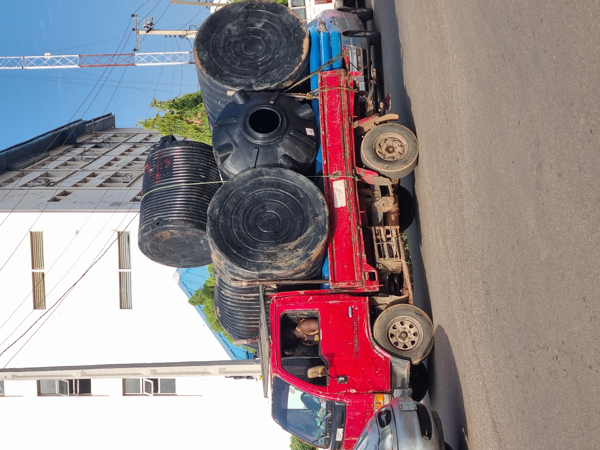 A red Nissan Cabstar truck laden with black water tanks is parked on a paved street in Jos, Nigeria. The truck is heavily loaded with five large water tanks. Adjacent to the truck are buildings, one white with rectangular windows and the other a taller structure with a visible communication tower. A silver-colored car is parked near the front of the truck. The scene is captured on a sunny day with a clear blue sky.