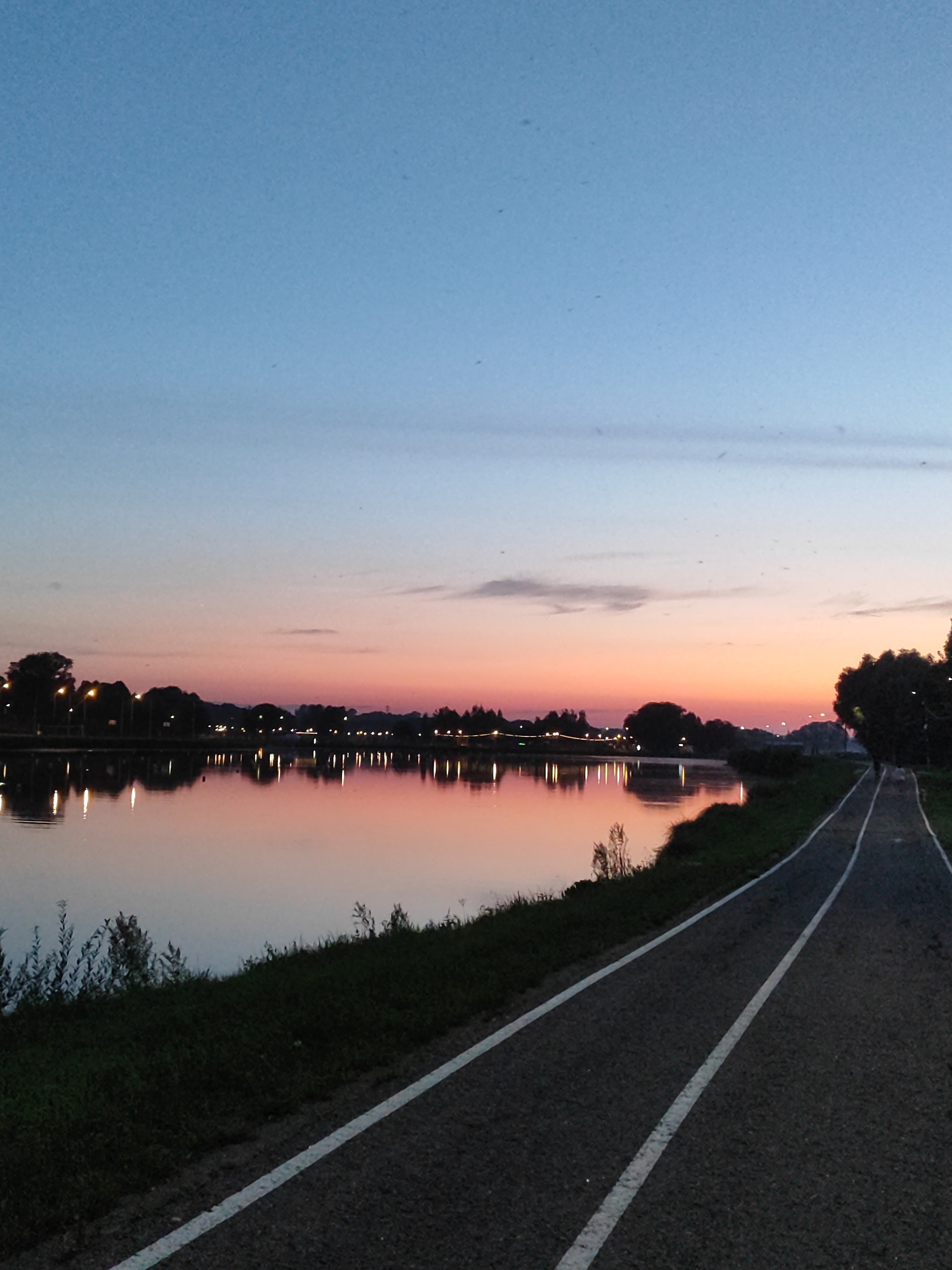 The image shows a sunset over a body of water, likely the Bronnitsky Pond in Bronnitsy, Russia. A paved path with white lane markings runs along the right side of the frame, bordered by grass and vegetation. Silhouetted trees and lights line the opposite bank of the water, with their reflections shimmering on the calm surface. The sky is a gradient of blue, pink, and orange, indicating the transition from day to night. There are no people visible in the frame.