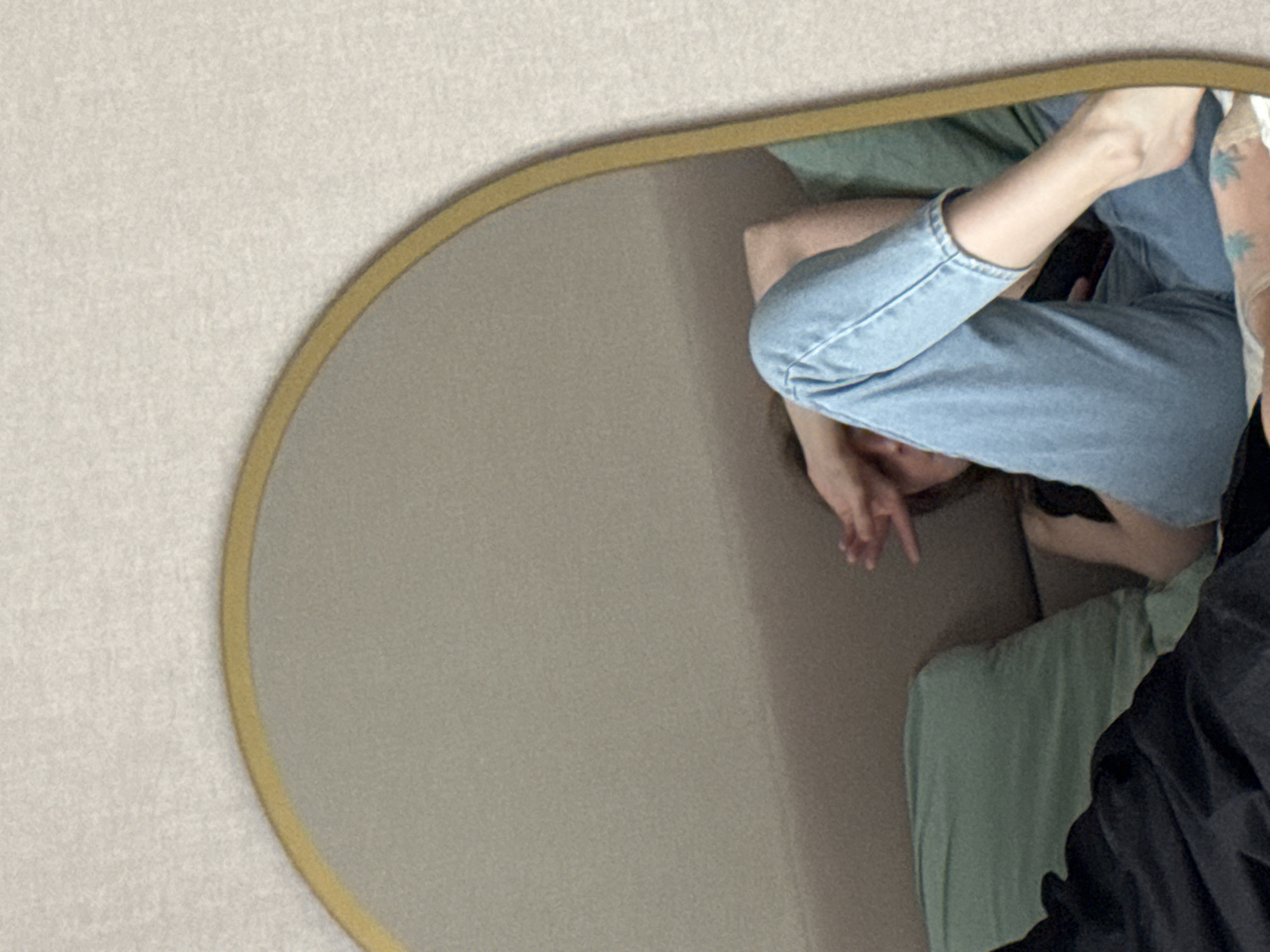 The image shows a mirror reflecting a person reclining on a bed. The person is wearing light blue jeans and a black top. The bed has a light green blanket or sheet. Part of a person wearing a black jacket is visible near the lower right corner of the mirror's frame. The surrounding wall is a neutral beige color. The city or country of origin cannot be determined from this image.