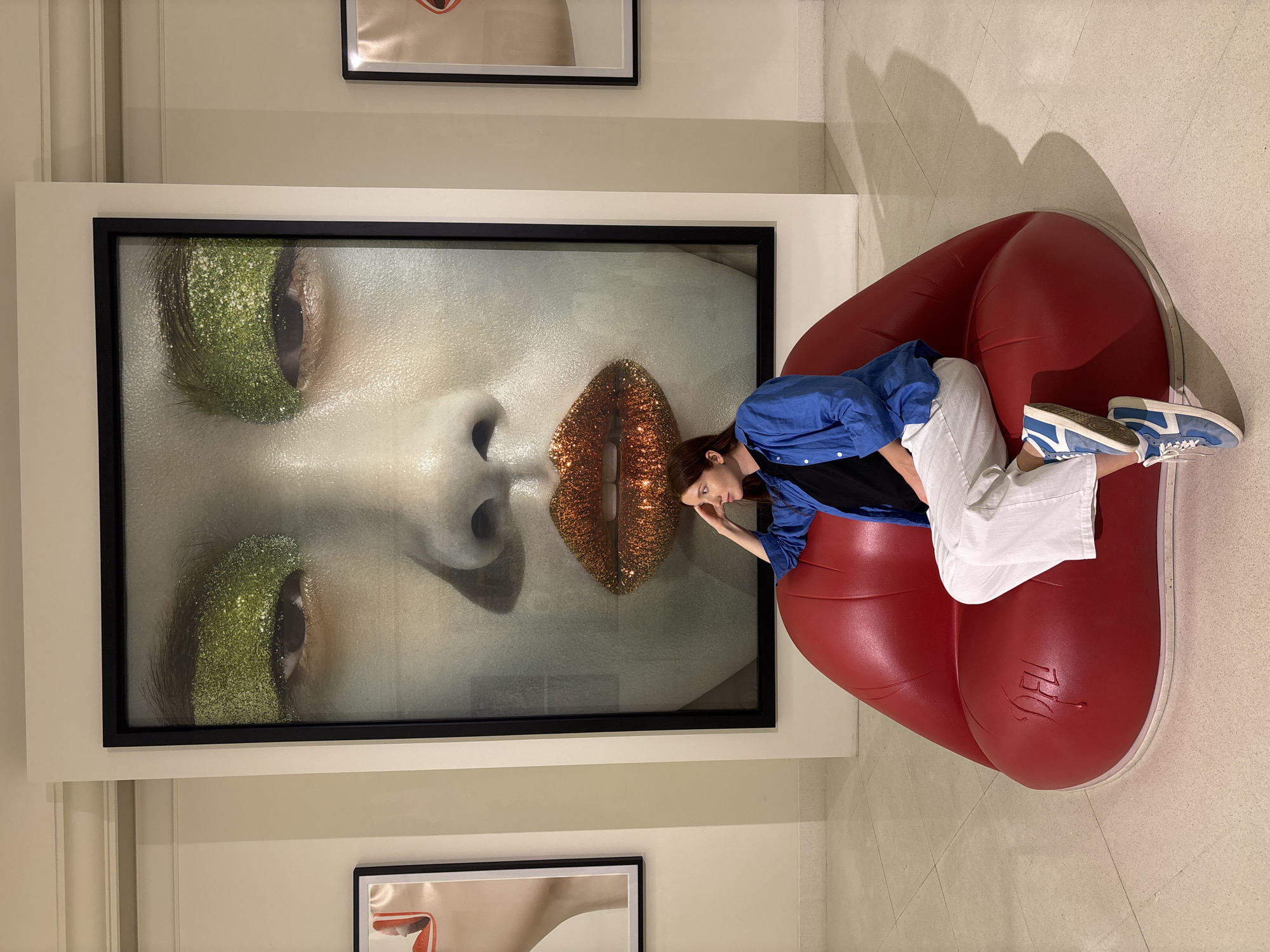 A full-length shot depicts a woman reclining on a red, lip-shaped sofa, positioned next to a large framed artwork of a woman's face. The artwork features close-up details of the woman's face, with heavy glitter eye makeup in green and sparkling orange lips. The woman reclining on the sofa is wearing white pants, a black shirt, and a blue collared shirt, and blue and white tennis shoes. The scene is set in a gallery or art space, evidenced by other framed artworks visible on the wall behind the sofa and the tiled floor. The sofa, the woman reclining on it, and the artwork are all centrally composed in the image. The sofa has the name "DELI" etched into it. It is likely located within Bangkok, Thailand.