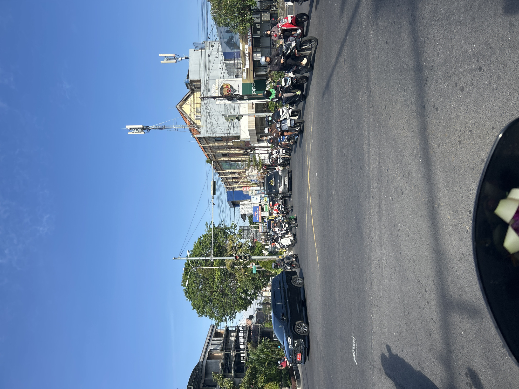 A street scene in Denpasar, Indonesia, shows a busy intersection with numerous motorcycles and a few cars navigating the road under a clear blue sky. The foreground is dominated by the asphalt of the road, with visible lane markings. A black SUV is prominent on the left, stopped or moving slowly. A line of motorcycles, each with a rider wearing a helmet, is proceeding through the intersection. Buildings line the sides of the street, displaying various signage, including "Kims Farma" and "PPO." Communication towers are visible on top of some buildings. A traffic light is present, indicating the intersection is controlled. The overall environment suggests a typical urban setting with moderate traffic flow during daylight hours.