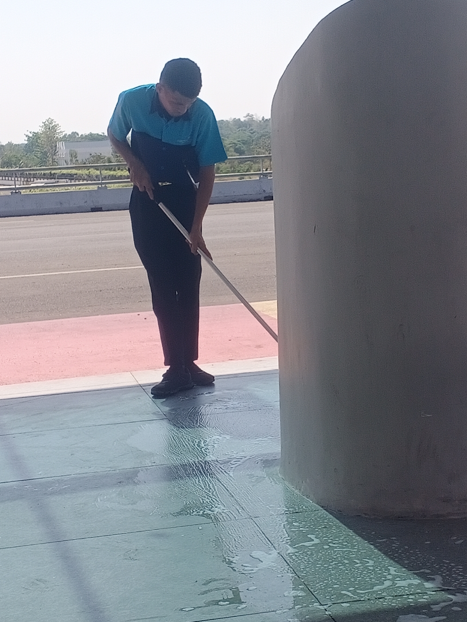In Baji Mangngai, Indonesia, a young person, likely male, wearing a two-tone blue and dark blue shirt and dark pants, is mopping a wet, tiled floor with a long-handled mop. The person is positioned with their back partially to the viewer, focusing on the task. The floor is visibly wet and soapy, with suds and streaks indicating recent cleaning. To the right of the person is a large, cylindrical, gray concrete pillar. In the background, beyond a pink and gray striped border, is an asphalt road and a walkway with some greenery and trees. The sky is clear and bright, suggesting daytime.