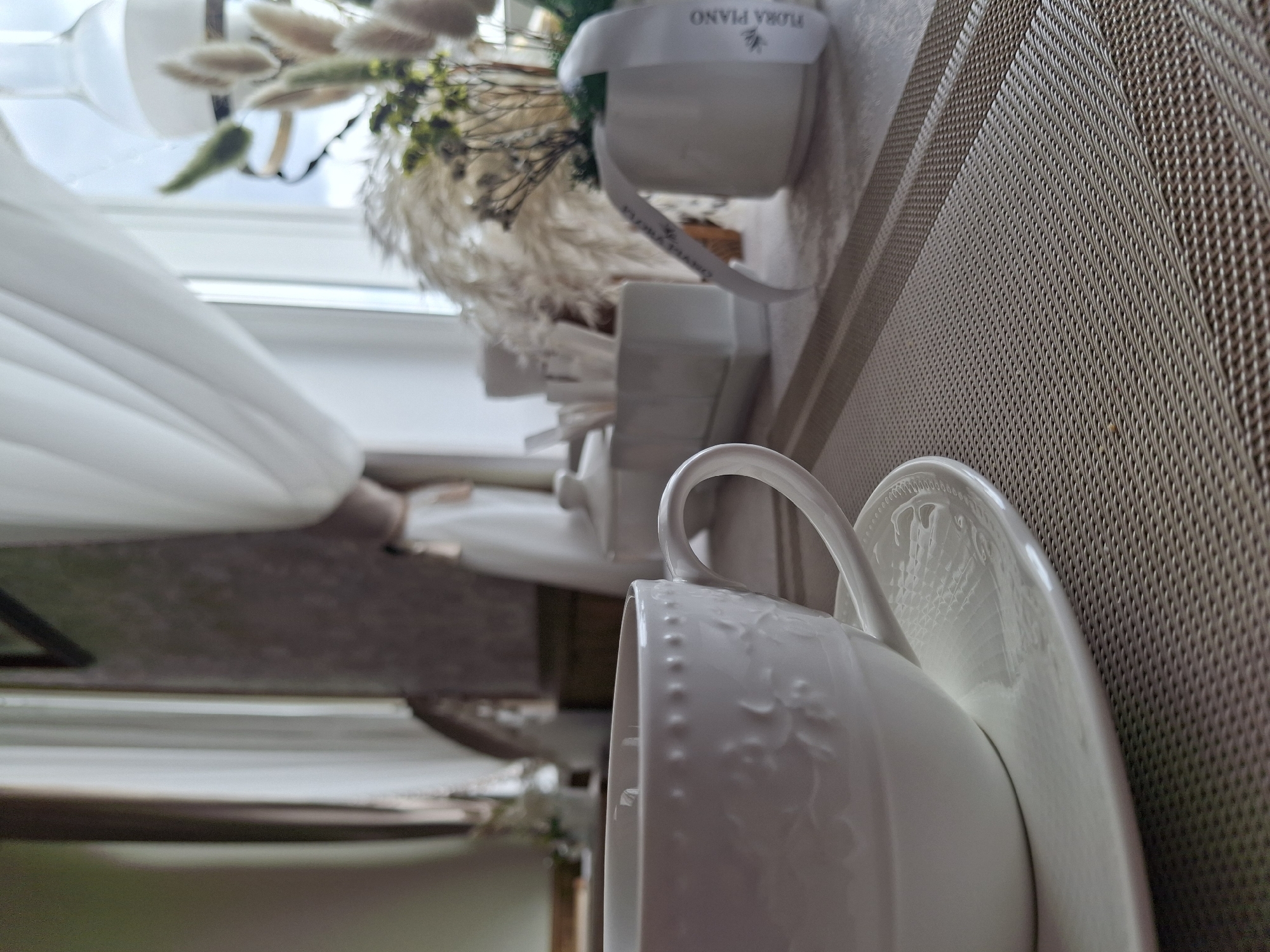 The image captures a close-up still life scene, likely indoors. In the foreground, a white teacup with a decorative, embossed pattern featuring floral motifs and a line of small raised dots near the rim is positioned on a matching white saucer. The cup's handle is prominent. Both the cup and saucer are resting on a light brown, textured placemat with a woven, mesh-like pattern.
In the mid-ground, slightly out of focus, a white ceramic container holds a dried floral arrangement. The arrangement consists of light, fluffy elements resembling pampas grass, mixed with some greener dried foliage and other delicate dried flowers. A white ribbon is tied around the ceramic container. Visible text on the ribbon, inverted, reads "FLORA PIANO" alongside a small insect or butterfly logo.
The background shows a window with white blinds or curtains, suggesting natural light is entering the room. The overall lighting is soft, indicating daytime. No specific activity is taking place; the scene portrays a calm and decorative arrangement. The setting appears to be a domestic space, possibly a table in a home or café.