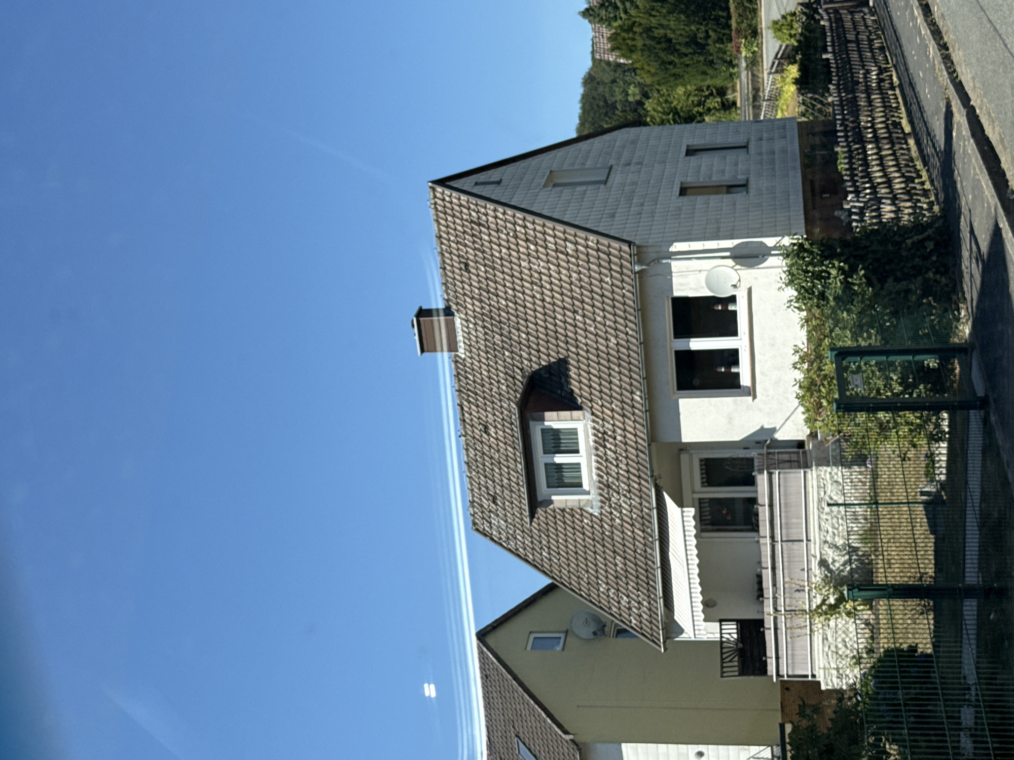 The image displays a residential area in Bad Sachsa, Germany, on a clear, sunny day. The foreground features a white house with a tiled roof, a front porch with an awning, and a fence. To the left, a portion of a yellow house is visible. The background shows trees and other residential buildings. The sky is a bright blue with faint contrails from aircraft. There are no people or animals visible in the scene. The scene appears to be a typical suburban street.