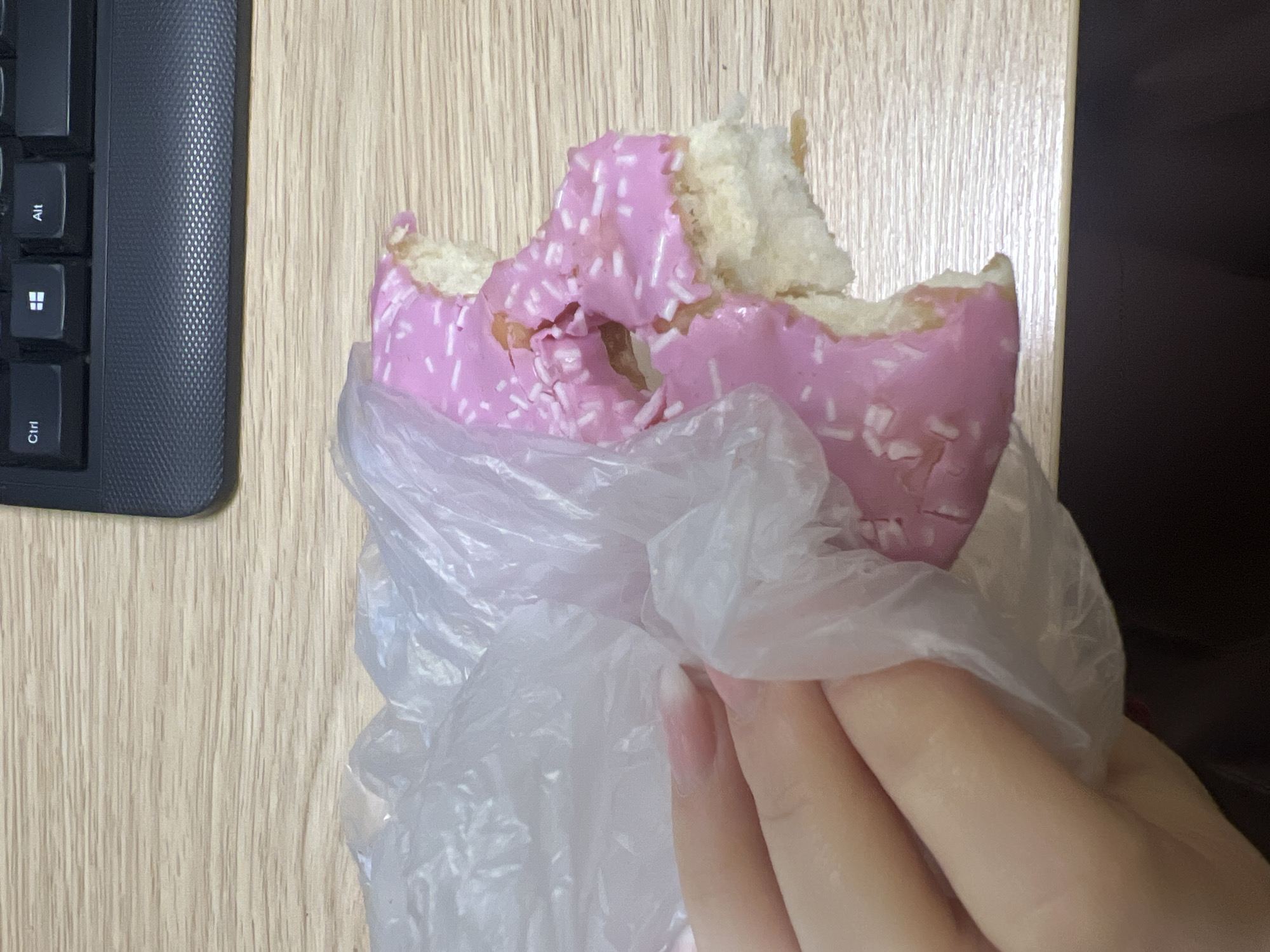 A partially eaten pink glazed donut with white sprinkles is held in a plastic bag. A hand holds the bag. A black keyboard is visible to the left. A wooden surface serves as the background. The location is unknown and cannot be determined from this image.