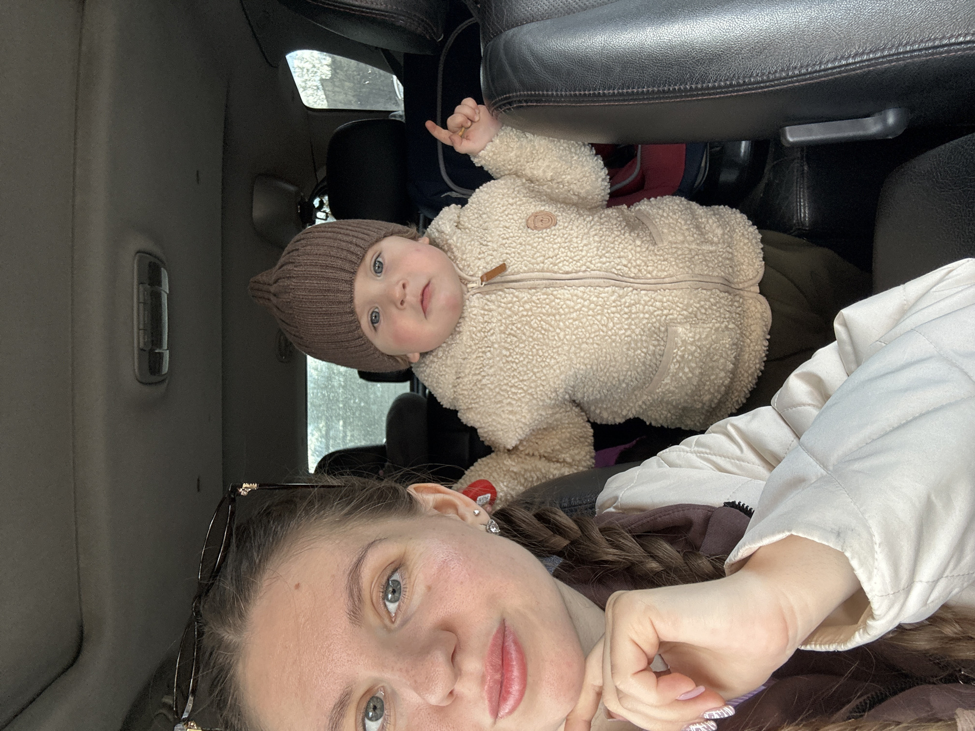 The image shows a selfie taken inside a vehicle, possibly a car. The photographer appears to be a woman with fair skin, light eyes, and brown hair styled in a braid. She is wearing glasses perched on her head and a light-colored jacket.
In the background, a baby wearing a brown knit hat and a cream-colored fleece jacket is visible. The baby is lying down, possibly in a car seat. The interior of the vehicle is visible, with dark leather seats and a window showing an indistinct, possibly urban, background.
The location is stated to be Klin, Russia, based on external information not visible in the photograph.