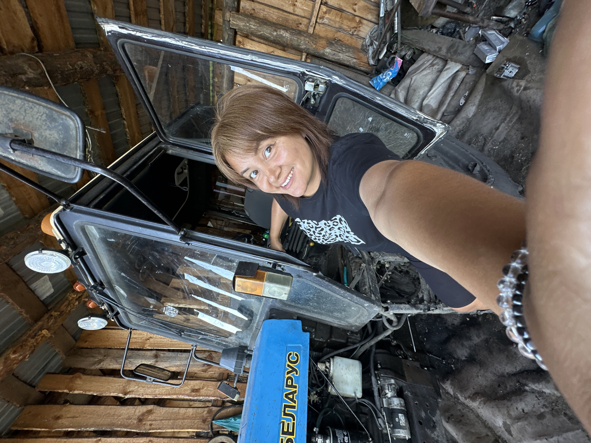 A woman with short brown hair is taking a selfie in a garage or workshop. She is wearing a black t-shirt with a white leopard print design. She is smiling and looking at the camera. The woman is leaning into the cab of a tractor, which appears to be under repair or maintenance. The tractor is dark gray with a transparent roof and doors. Parts of the engine and machinery are visible. The background shows wooden walls and ceiling, and various tools and materials are scattered around. A blue box with yellow Cyrillic text "БЕЛАРУСЬ" (Belarus) is visible on the tractor. The lighting suggests it is daytime, possibly with natural light filtering in. The overall scene conveys a sense of casual work in progress.