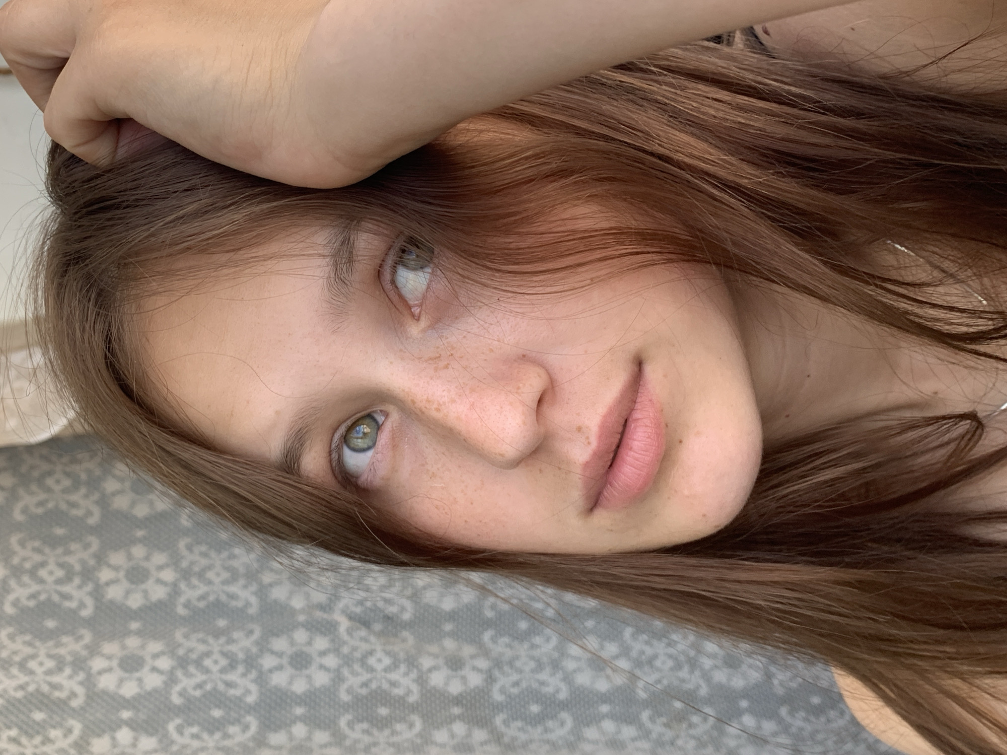 A close-up portrait shows a young woman with long, brown hair and green eyes, her hand resting on her head. She has a light complexion with freckles across her nose and cheeks. Her lips are slightly parted, and she appears to be looking away from the camera. The background is a textured light blue wallpaper with a white floral pattern. The lighting suggests an indoor setting, possibly a home, and the overall atmosphere is casual and natural. The image is cropped to focus on the woman's face and upper torso. The location is not visible in the image.