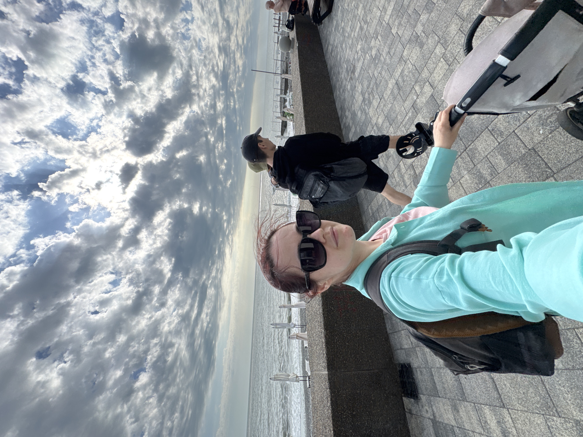 A woman with sunglasses, wearing a turquoise hoodie and a backpack, is pushing a stroller along a paved walkway in Sochi, Russia. She is oriented towards the camera. A man in a black sweatshirt and baseball cap is walking nearby. The background shows the sea with a row of closed umbrellas and a cloudy sky. The location is a promenade near the beach.