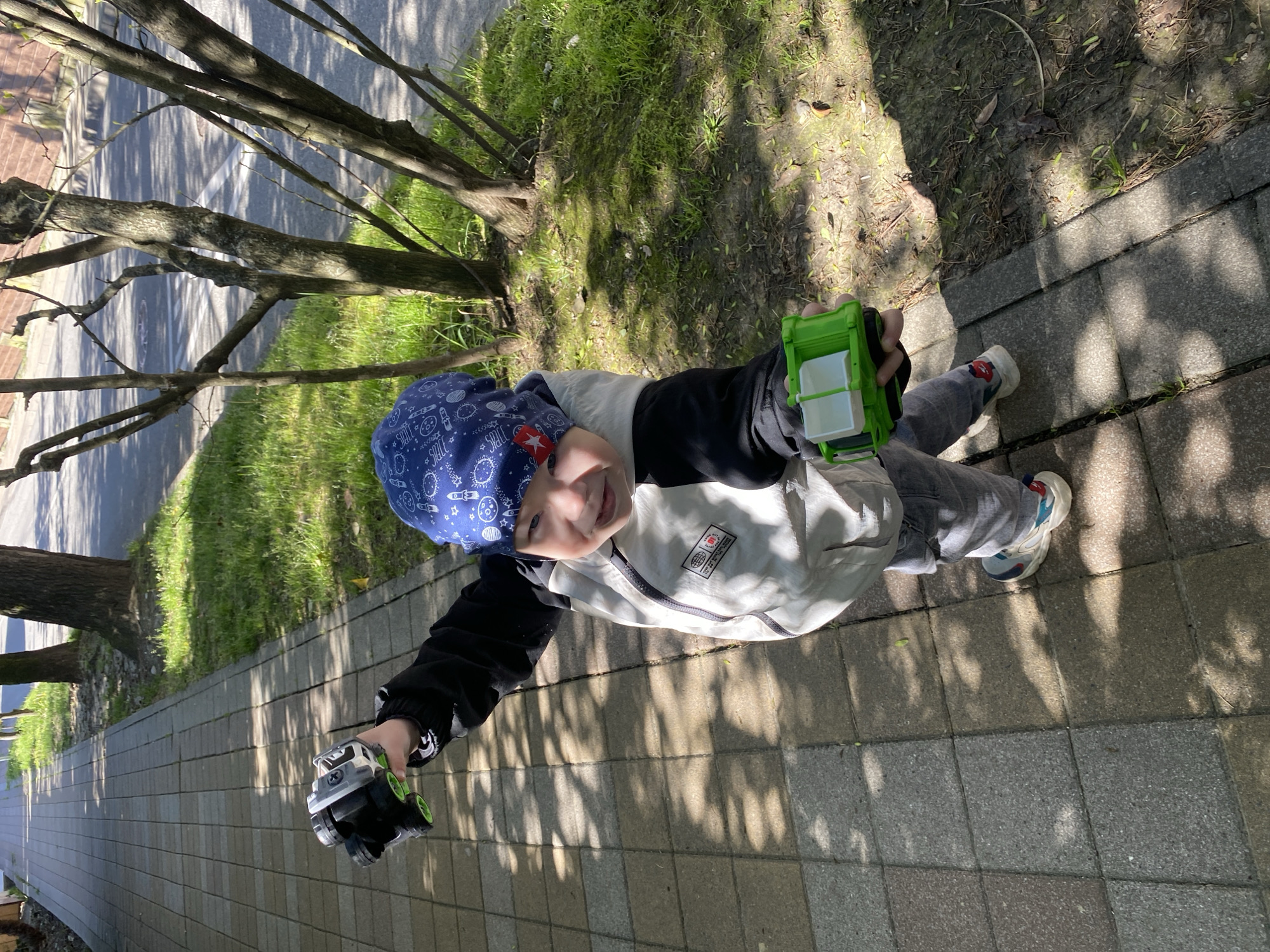 The media file, presented on its side, captures a young child from an overhead perspective, smiling towards the camera. The child is positioned in the lower-left quadrant of the frame, standing on a grey tiled path that runs vertically along the right side of the image.
The subject is a boy, wearing a blue beanie adorned with white space-themed patterns including rockets, planets, and the word "SPACE." He wears a two-tone jacket, light grey or white on the torso and dark on the sleeves, over grey pants. His sneakers are white with blue and red details. In his left hand (relative to his body), he holds a small grey and green toy truck. In his right hand, he holds a bright green toy garbage truck with white compartments.
The setting is an outdoor area in Sochi, Russia, likely a park or a sidewalk next to a street. To the left of the child and the path is a strip of green grass and patches of dirt. Beyond the grass, several trees with mostly bare branches rise, casting prominent, sharp shadows across the ground and path, indicating a sunny day, likely late morning or early afternoon. A paved road with faint white markings is visible in the distance through the tree branches. The overall mood is cheerful, with the child appearing happy and engaged.