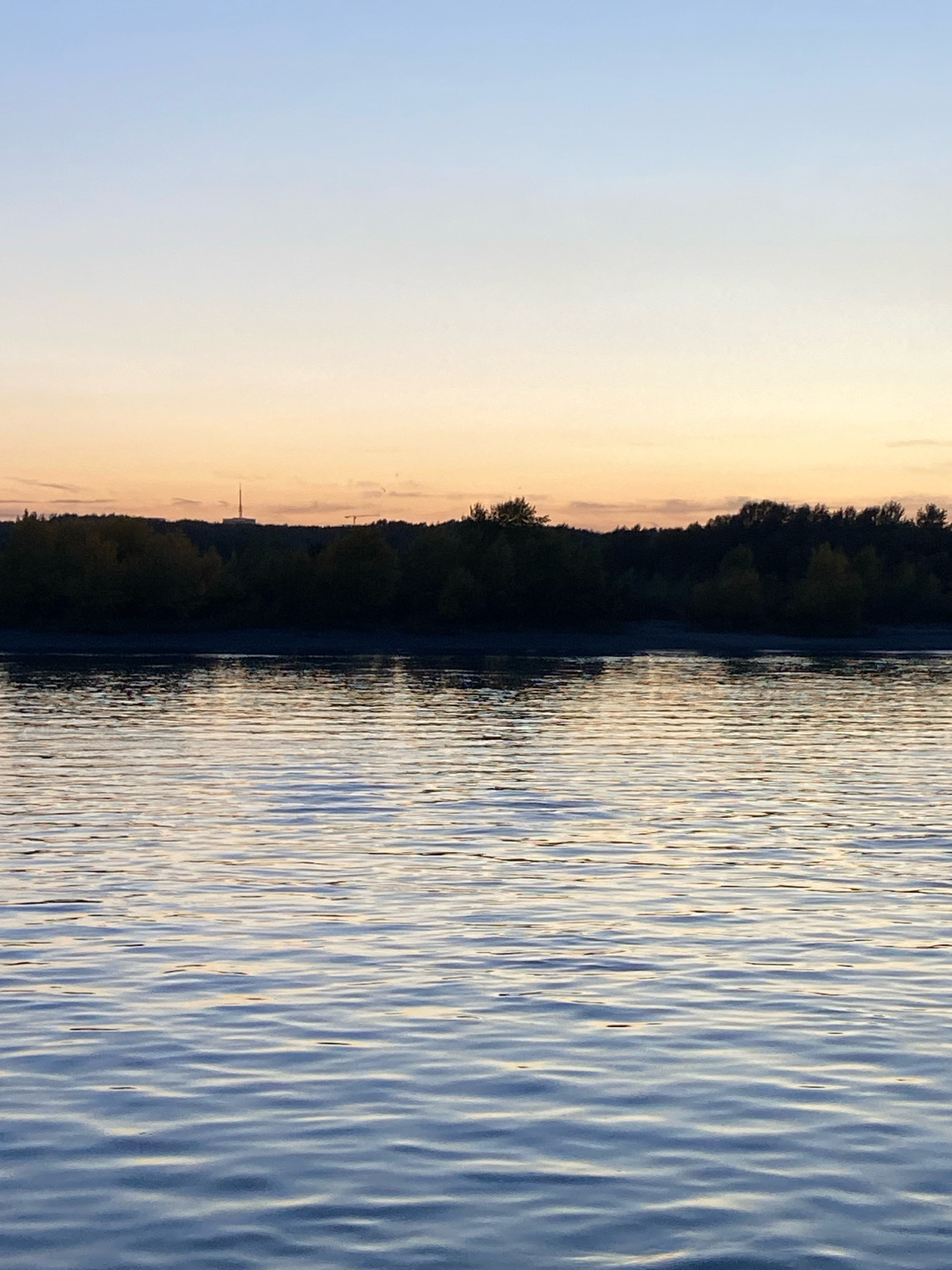 The image depicts a serene sunset over the Ob River in Novosibirsk, Russia. The sky transitions from a pale blue at the top to warm hues of orange and yellow near the horizon. A dark, silhouetted treeline stretches across the middle ground, with a prominent radio tower visible above the trees in the distance, suggesting a more urbanized area beyond the natural landscape. The river occupies the foreground, its surface rippled by gentle waves that reflect the colors of the fading sky. No people or active events are present; the scene is characterized by its tranquil, natural beauty.