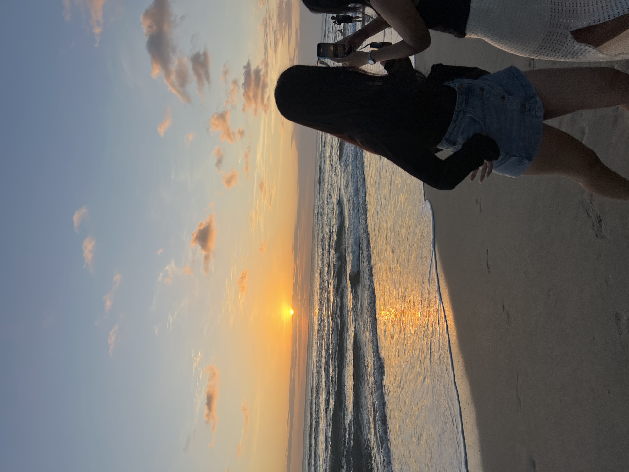 The image depicts a beach scene in Kuta, Indonesia, during sunset. The sun is low on the horizon, casting a warm orange glow over the ocean and the sky. The waves are gently rolling onto the shore, creating a foamy white line where they meet the sand. Two women are standing on the beach, facing the ocean. The woman in the foreground is wearing a black long-sleeved top and a denim skirt. She has long dark hair and is posing with her hand on her hip. The woman next to her is wearing a black top and a white skirt. She is holding up her phone and taking a picture of the sunset. In the background, other people can be seen walking along the beach. The sand is a light brown color, and there are footprints visible in the sand. The sky is a gradient of blue and orange, with some scattered clouds. The overall atmosphere is serene and picturesque, capturing the beauty of a tropical sunset.