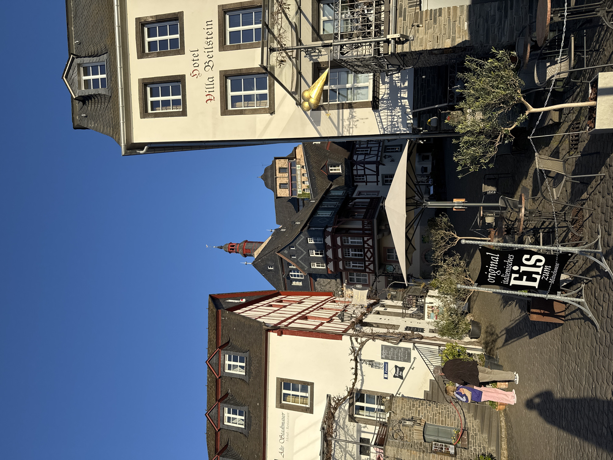 The image shows a street scene in Beilstein, Germany, with several buildings, signage, and people.
Buildings: There are at least three buildings visible. The building at the top, labeled "Hotel Villa Beilstein," has a light-colored facade and is several stories high. A second building has a half-timbered style with a white facade and red accents. A sign on the building reads "Alte Stadtmauer Hotel - Restaurant" and "Zimmer frei." A third building is visible in the background.
Signage: There are multiple signs, including "Hotel Villa Beilstein," "Alte Stadtmauer Hotel - Restaurant," and a sign that reads "original italienisches Eis zum Mitnehmen."
People: Two people, an adult and a child, are walking on the brick-paved street/sidewalk. The adult is wearing a dark jacket and light pants, and the child is wearing a pink and blue outfit.
Objects: There is outdoor seating associated with the businesses, including tables, chairs, and umbrellas. A potted tree and some decorative foliage are also visible.