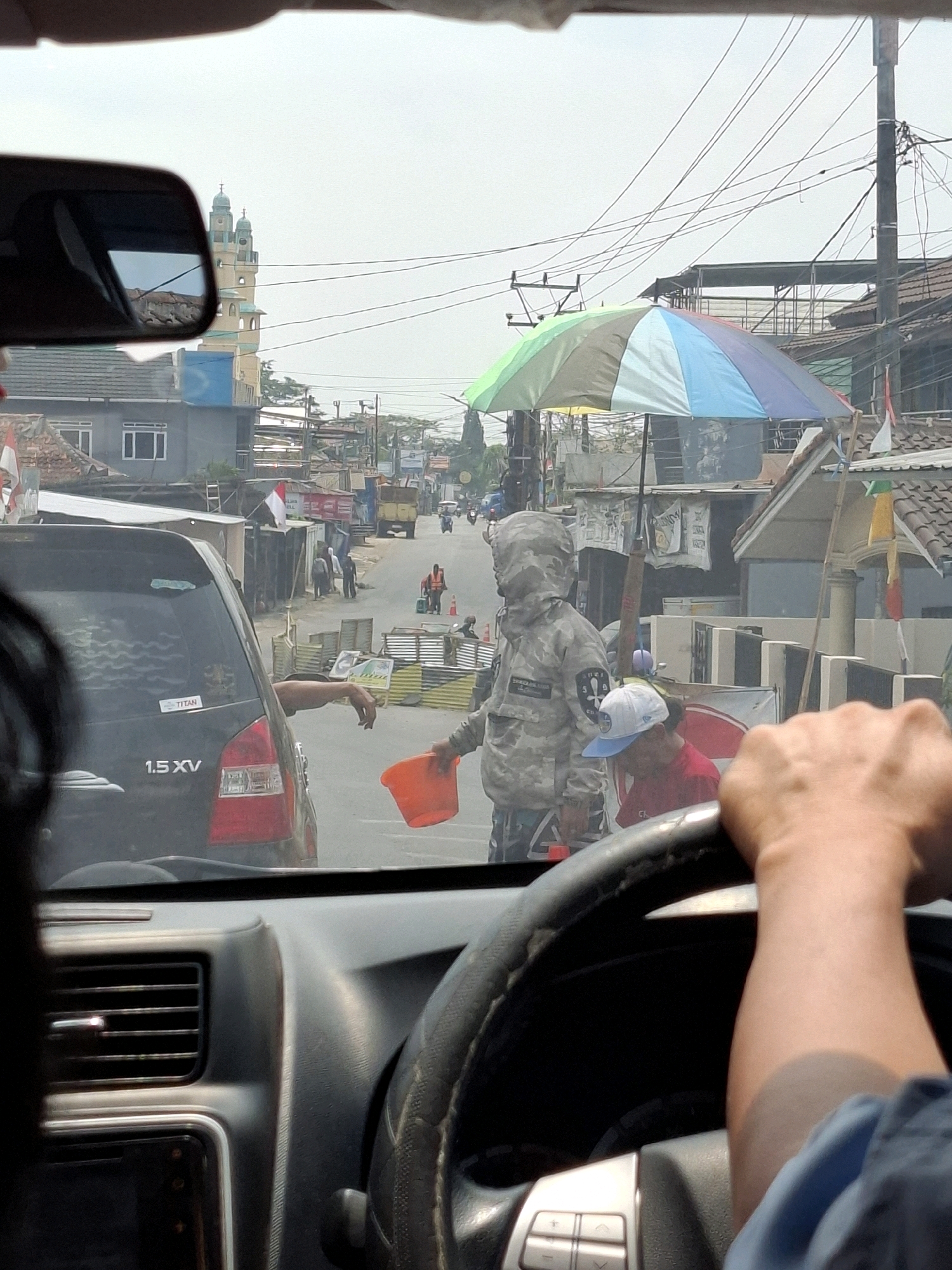 The image shows a view from inside a vehicle looking forward towards a street scene in Sukabumi, Indonesia. The driver's hands are visible on the steering wheel. Outside the vehicle, on the right side of the road, a person in a camouflage jacket and a person wearing a red shirt and a blue and white cap are interacting. The person in the camouflage jacket is holding an orange bucket, and appears to be receiving something or interacting with the person in the red shirt. A multi-colored umbrella is set up nearby, providing shade. The road is lined with buildings and structures typical of a city environment, with power lines crisscrossing overhead. A black SUV is visible in front of the viewer's vehicle. In the distance, a mosque with two minarets can be seen. The overall impression is a busy street scene, possibly a market or intersection, with pedestrians and vehicles.