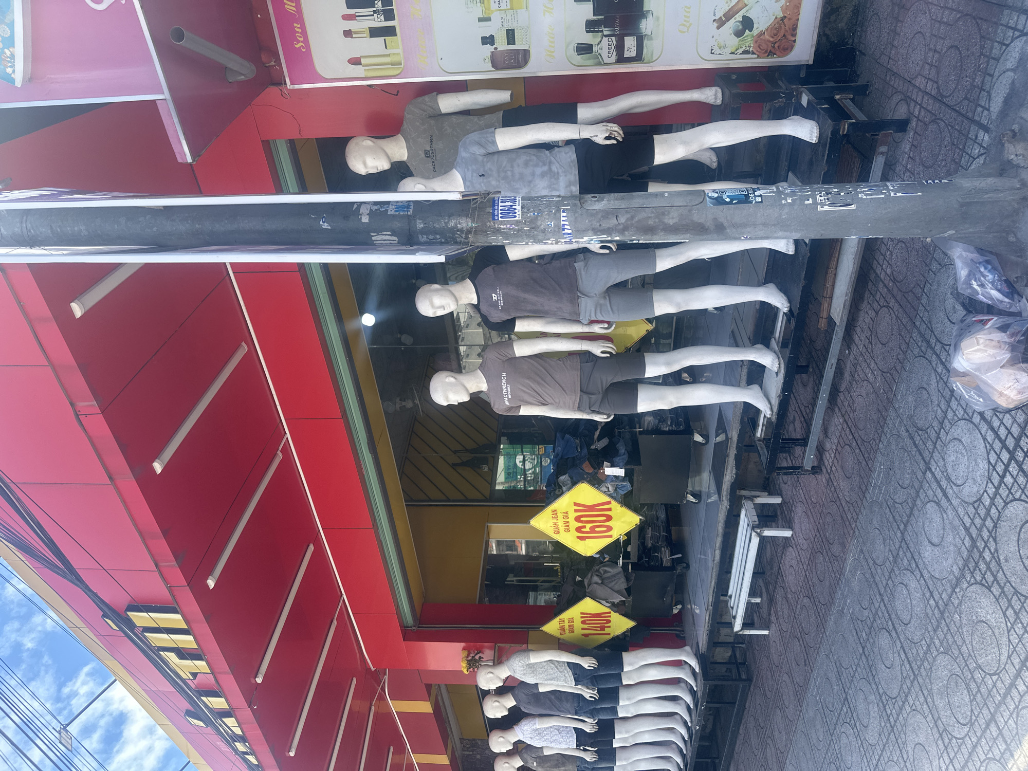 A street-level, medium shot captures the storefront of a clothing retailer in Nha Trang, Vietnam. The building facade is painted a vibrant red with yellow accents on the signage, which appears to read "QUAN TI" in large gold letters, possibly indicating the store's name. Underneath the prominent red awning, several mannequins are displayed outside, dressed in casual men's apparel, primarily t-shirts and shorts. Two prominent yellow diamond-shaped signs with black text are visible, advertising prices: one reads "QUAN TAY GIA GIAM 140K" and the other "QUAN JEAN GIA GIA 160K," suggesting discounted prices for trousers and jeans respectively. Behind the mannequins, the store's interior is visible through glass doors and windows, showing racks of clothing. A utility pole, covered in stickers and tape, stands directly in front of the display, partially obscuring the view of some mannequins. The sidewalk in front of the store is paved with patterned concrete tiles. Plastic bags containing items are scattered on the ground near the base of the utility pole. The overall scene depicts a commercial street environment with a focus on clothing retail and ongoing sales promotions.