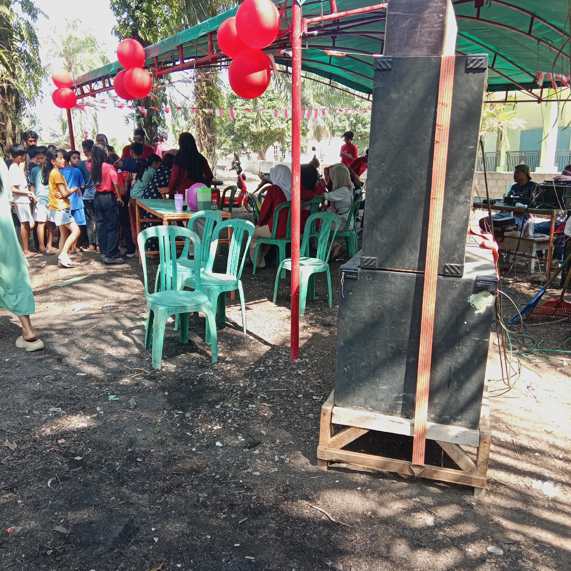 A group of people, mostly children and teenagers, are gathered outdoors under a green awning supported by red metal poles. Red balloons are strung above. Several green plastic chairs are set up, some empty and some occupied by individuals. In the foreground, a large black speaker system is mounted on a wooden structure. A red and orange strap is attached to the speaker. In the background, there are trees and some buildings, including a light-colored wall. The ground is a mix of dirt and scattered debris. The scene appears to be set up for an outdoor event or gathering. The presence of Indonesian flags, visible in the cropped images, suggests the event may be related to Indonesian Independence Day, which is celebrated on August 17th. Therefore, this scene is likely in Dumai City, Indonesia, during a local celebration.