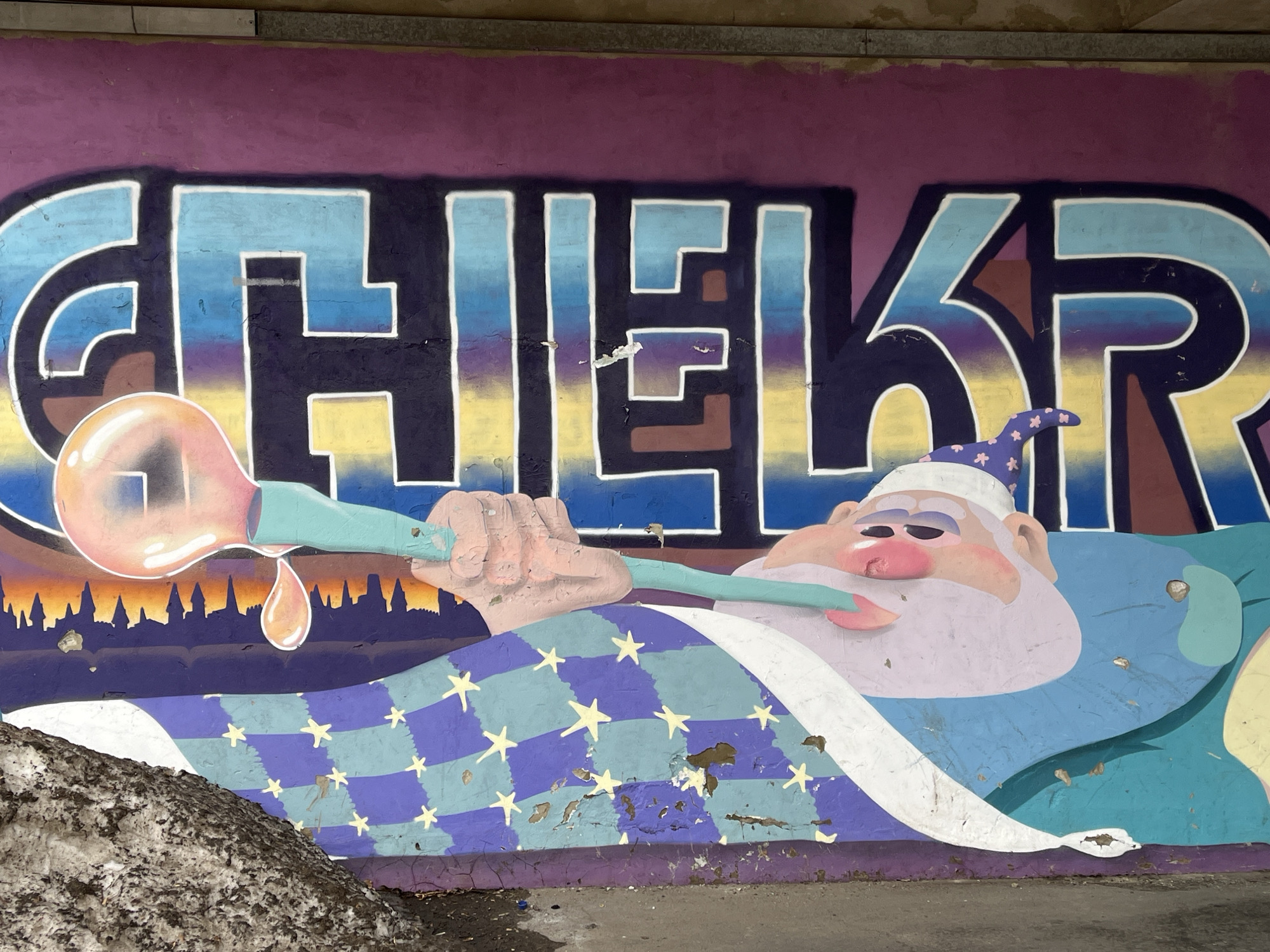 A colorful graffiti mural occupies a weathered concrete wall, likely beneath an underpass or on a retaining wall, in Krasnoyarsk, Russia. The mural features the stylized block letters "CHILLKR" (or "CHILLKР") with a blue, purple, and yellow gradient fill and white outlines, set against a solid purple background. Below the text, a cartoon-style figure resembling a sleeping wizard is depicted. The wizard has light pink skin, a prominent pink nose, a white beard, and wears a white pointed cap adorned with blue stars. He is clad in light blue and covered by a blanket patterned with alternating purple and light blue stripes and yellow stars. The wizard holds a light blue pipe-like object with a glowing pink-orange bulb at its end, from which a single pink-orange drop is suspended. Behind the wizard's head, a dark silhouette of a city skyline is visible against an orange and purple gradient sky. The concrete wall exhibits considerable wear, including paint chipping and minor damage, particularly on the lower sections of the artwork. The ground directly in front of the mural is a dirty grey concrete or asphalt surface. A pile of dark, slushy snow and debris is present in the lower-left corner, partially obscuring the blanket portion of the mural.