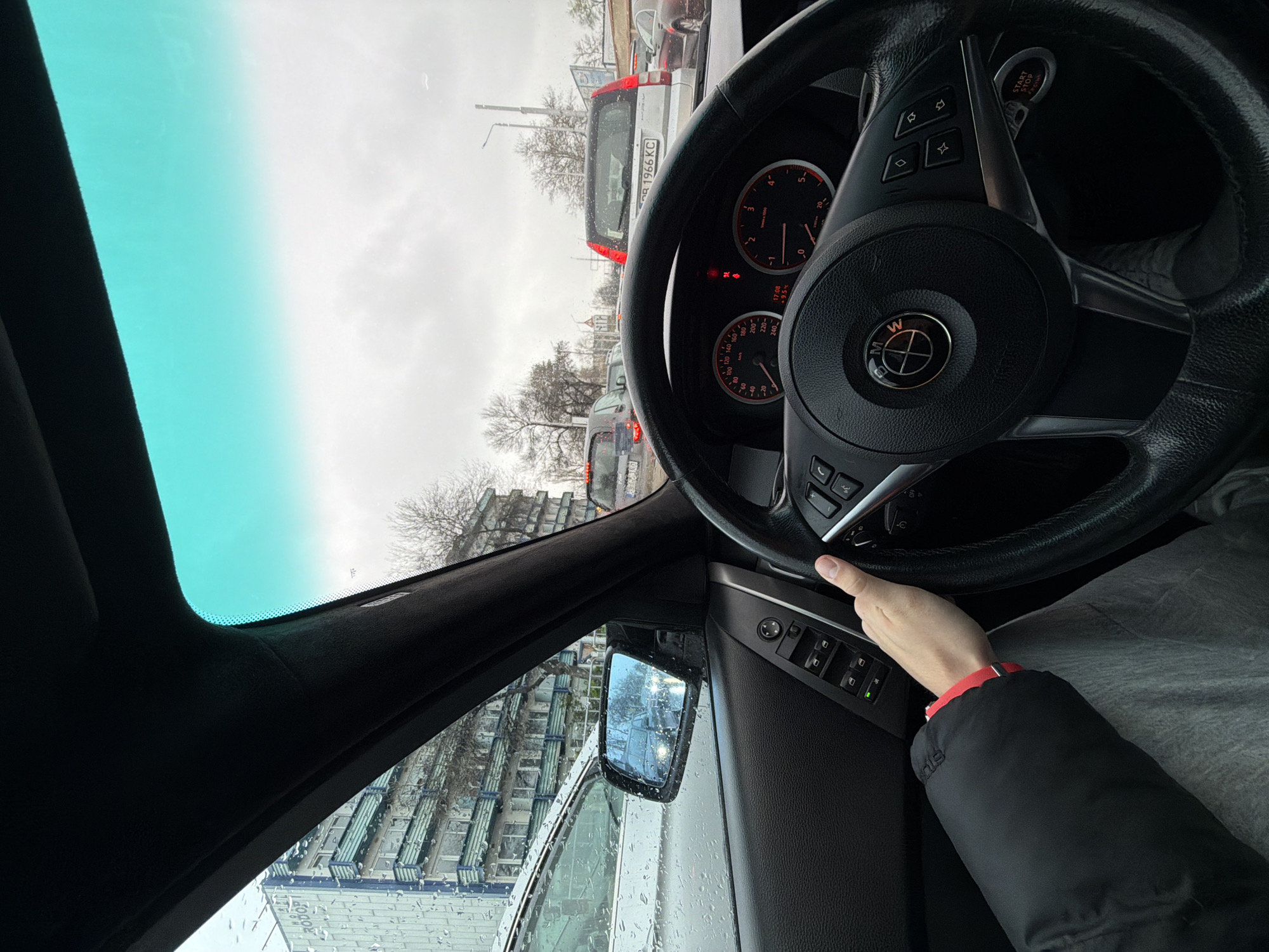 The image shows the view from inside a car, likely a BMW, with the steering wheel and dashboard prominently displayed. The driver's hand rests on the steering wheel. Outside the car, other vehicles are visible, suggesting traffic. The vehicle in front has a license plate "PB 1966 KC." The background includes buildings and trees, indicating an urban environment. The side mirror reflects more of the city landscape. Raindrops are visible on the car window. The time on the dashboard is 17:08 and the fuel level is at 9.5%. Based on the license plate, the location is the city of Plovdiv, Bulgaria.