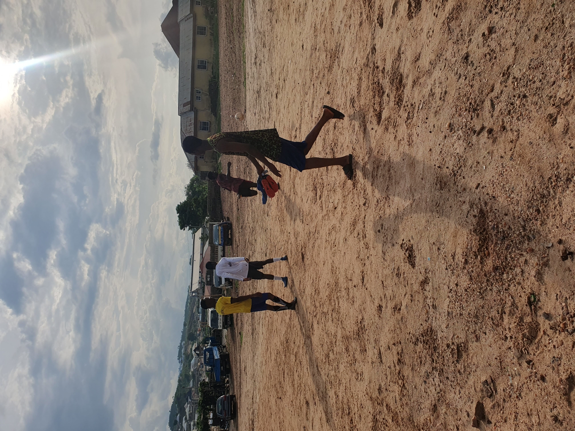 The image depicts an outdoor scene in Lokoja, Nigeria. The setting appears to be a sandy field or vacant lot. The sky is partially cloudy with the sun shining through.
Several individuals are visible, mostly young men. One is running with a red and blue object in his hand. Others are standing nearby, some wearing yellow or white shirts.
In the background, there are buildings, trees, and parked vehicles. A two-story building with a brown roof is partially visible. Further in the distance, the terrain slopes downward towards what might be the outskirts of the city.