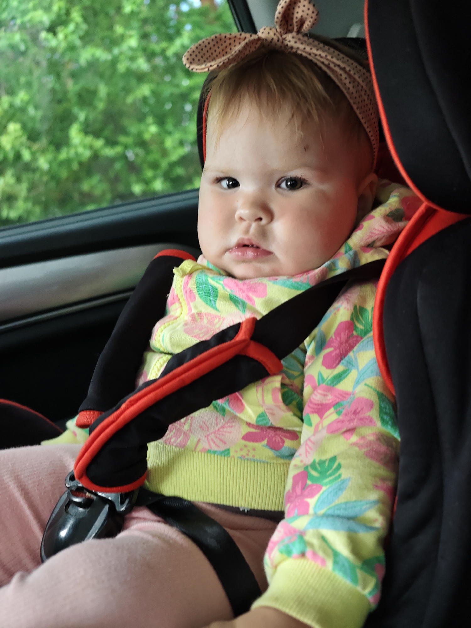 A young child, identified as a baby, is depicted seated in a car seat within a vehicle, located in the city of Chaikovsky, Russia. The child has fair skin and light brown hair, wearing a light brown, polka-dotted headband tied into a bow at the top of the head. The child's dark eyes are directed towards the viewer, maintaining a neutral expression. The baby is dressed in a long-sleeved top with a yellow base, featuring a vibrant floral and leaf pattern in pink, green, and light blue, paired with light pink pants. The child is secured in a black car seat with prominent red accents outlining the harness straps and padding. A five-point harness system is visible, with black straps extending over the shoulders and across the chest, converging into a black buckle assembly positioned between the child's legs. Through the car window in the background, out-of-focus green foliage, indicative of trees or bushes, is visible under daylight conditions.