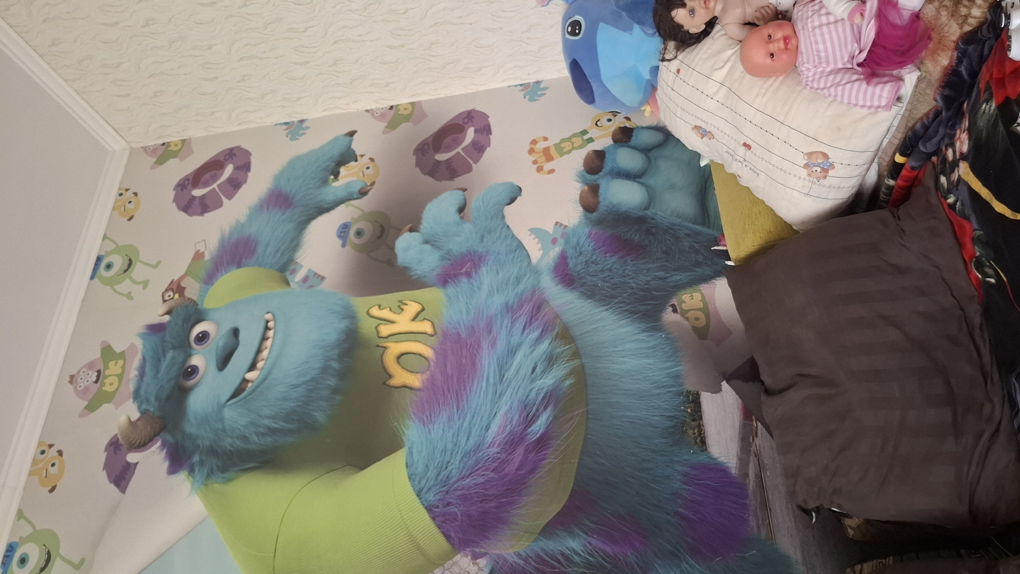 The image depicts an indoor scene, likely a child's bedroom, featuring a large mural of the character Sully from Monsters, Inc. on the wall. The mural covers a significant portion of the wall, showing Sully in his signature green shirt with the word "OK" on it, and his blue and purple fur. The background of the mural is adorned with various cartoon monsters. In the foreground, several plush toys and pillows are visible. These include a blue Stitch plush toy, a doll with brown hair, and a baby doll wearing a striped shirt and a pink skirt. A dark brown striped pillow and a yellow pillow are also present, along with a colorful blanket. The lighting suggests it is daytime, with natural light potentially coming from an unseen window. There is no indication of the specific location within the city of Shymkent, Kazakhstan, as the image focuses on the interior decor and objects.