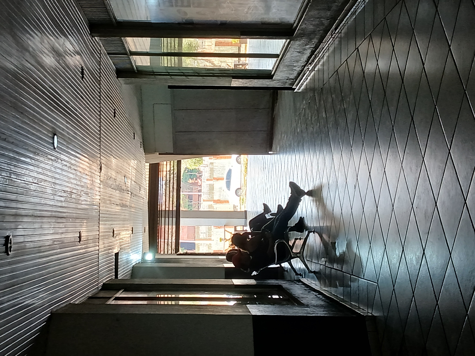 This image captures an indoor-outdoor transition area, likely a hallway or waiting space, in Ciudad Guayana, Venezuela, under bright daylight. The perspective is from an elevated position, looking down into the corridor.
On the left, a wall is covered in horizontal, corrugated metal panels, featuring several small, circular lights. Opposite this, the floor and an adjacent wall are clad in dark gray tiles arranged in a distinct diamond pattern. These tiles reflect the strong sunlight entering the space.
At the far end of the corridor, intense natural light pours in from an open doorway or large window, creating a bright area on the floor and casting long, sharp shadows. Through this opening, elements of an outdoor scene are visible: green foliage, parts of buildings, and a white car, indicating an urban or suburban setting.
Two individuals are seated within this brightly lit zone. They appear in silhouette against the strong backlight, resting on what seems to be a single office-style chair with wheels. One person is clearly visible, facing away from the camera, with their feet propped up in a relaxed posture, suggesting they are waiting or resting. A second individual or an object is less distinctly visible near the first.
In the foreground, dark, angular concrete structures or steps are present, adding to the architectural composition. The time of day appears to be sunny, possibly late morning or early afternoon, given the intensity and direction of the light. There is no visible text in the image. The scene conveys a sense of quiet and repose within a structured environment.