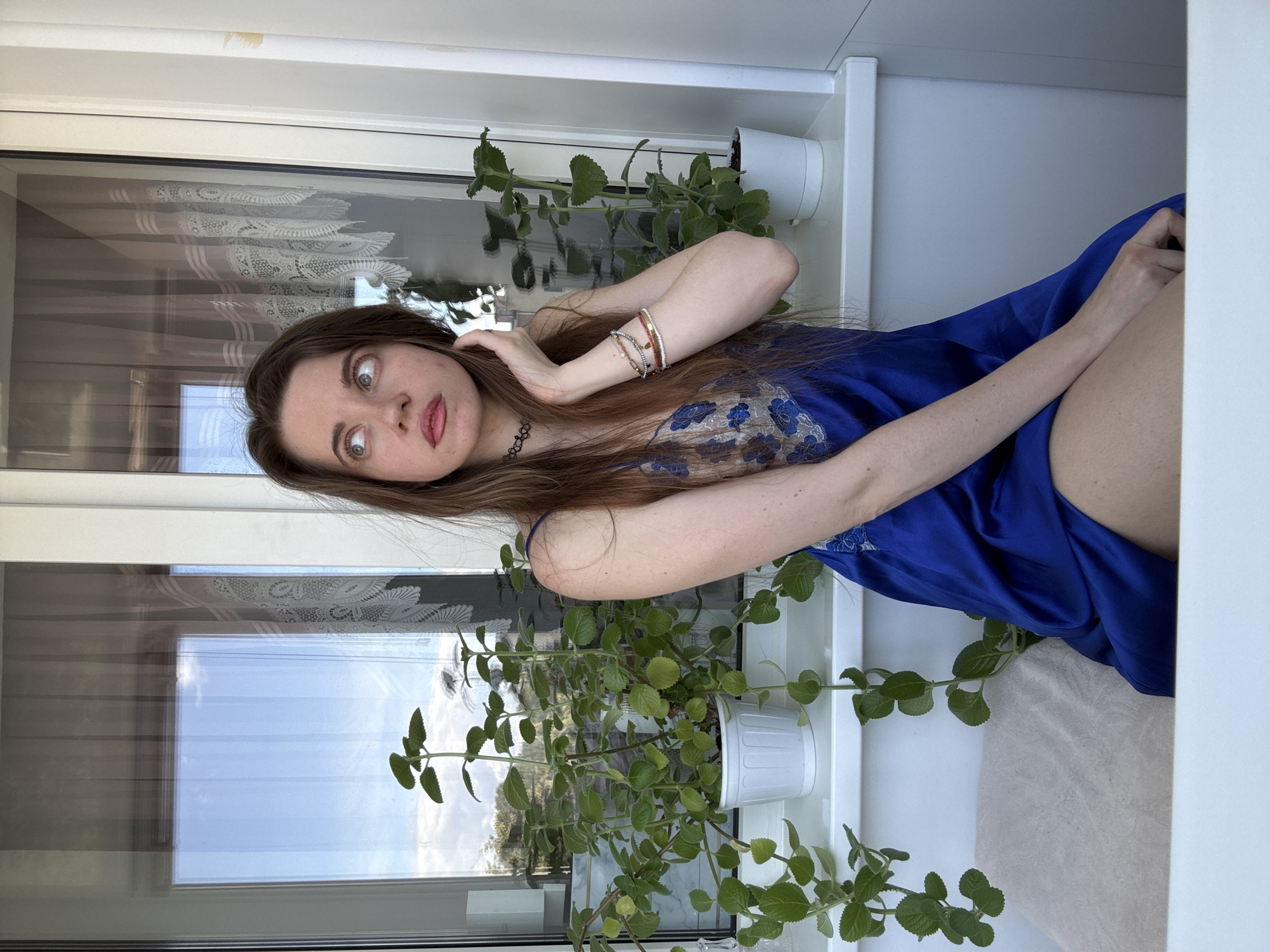 A young woman with long brown hair and wide blue eyes is posed indoors, leaning against a window frame. She is wearing a blue satin slip with lace detailing on the bust and a black choker necklace. Several bracelets adorn her left wrist. She is looking towards the camera with an expressive, slightly surprised gaze.
The scene is set on a windowsill with two potted green plants on either side of the woman. The window behind her is paneled, with sheer curtains that have a lace pattern. Outside the window, the sky appears bright blue with some clouds, suggesting daytime and fair weather. The room's interior is white and minimalist. The overall impression is of a casual, perhaps intimate, moment captured in soft natural light. There is no visible text in the image.