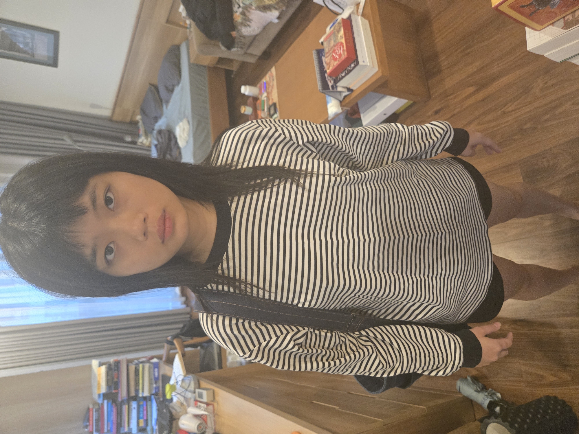A young Asian woman stands in a room with hardwood floors. She wears a black and white striped long-sleeved shirt, black shorts, and has a black strap across her body. The room contains a coffee table with books, a sofa, a bookshelf filled with books, and a window with blinds. The location is likely in a residential area of Hà Nội, Vietnam.