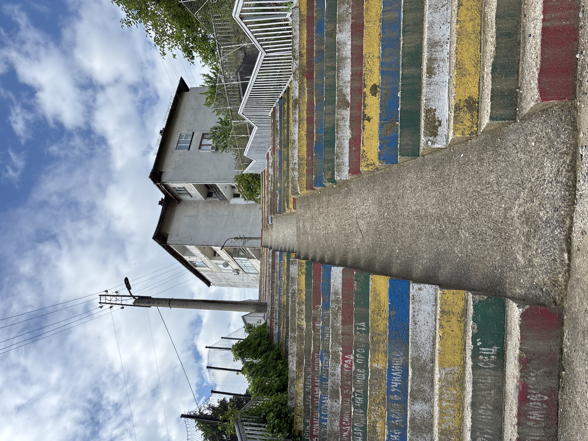 Here's a detailed description of the image:
The image showcases a long flight of concrete stairs in Zlatograd, Bulgaria, distinguished by their vibrant, alternating colors of yellow, blue, white, red, and green on the risers. Inscriptions are etched into the painted risers of the stairs. To the left of the stairs, a weathered, grey building stands against a blue sky dotted with white clouds.
A concrete utility pole with power lines is visible beside the building. A chain-link fence runs along the base of the stairs, partially obscured by greenery. On the right, the bare concrete tread of the stairs provides a pedestrian walkway alongside the colored risers. Above, a white railing accompanies the stairs, suggesting they ascend a significant height.