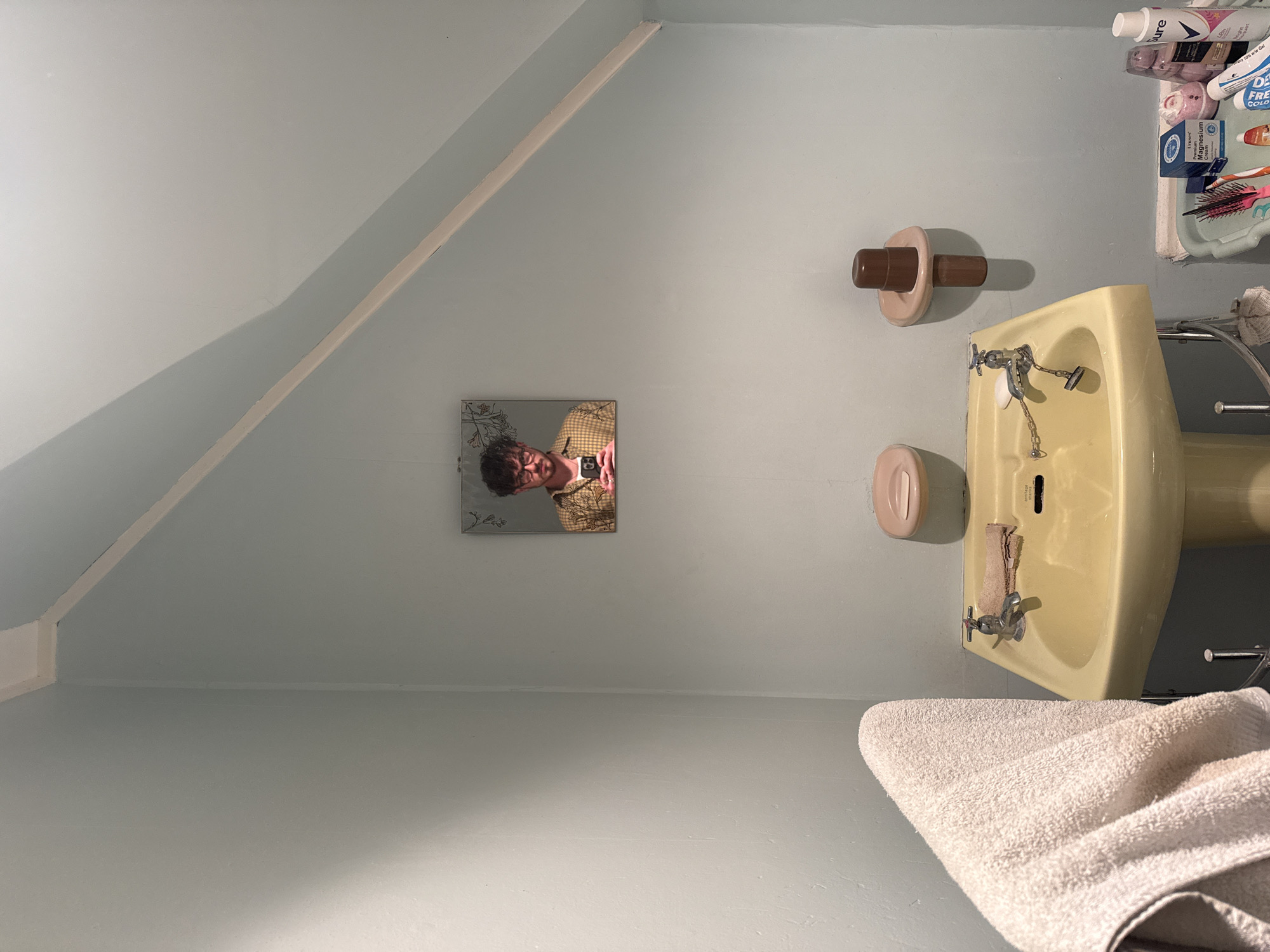 The image shows a bathroom interior with light blue walls and a sloping ceiling. A yellow sink with silver faucets is positioned to the right, accompanied by a brown and tan toilet paper holder above and a pink soap dish below. A white towel hangs near the bottom of the frame. Above the sink, a shelf holds various toiletries. A square mirror on the left wall reflects a person wearing glasses and a yellow patterned shirt. The location is unknown.