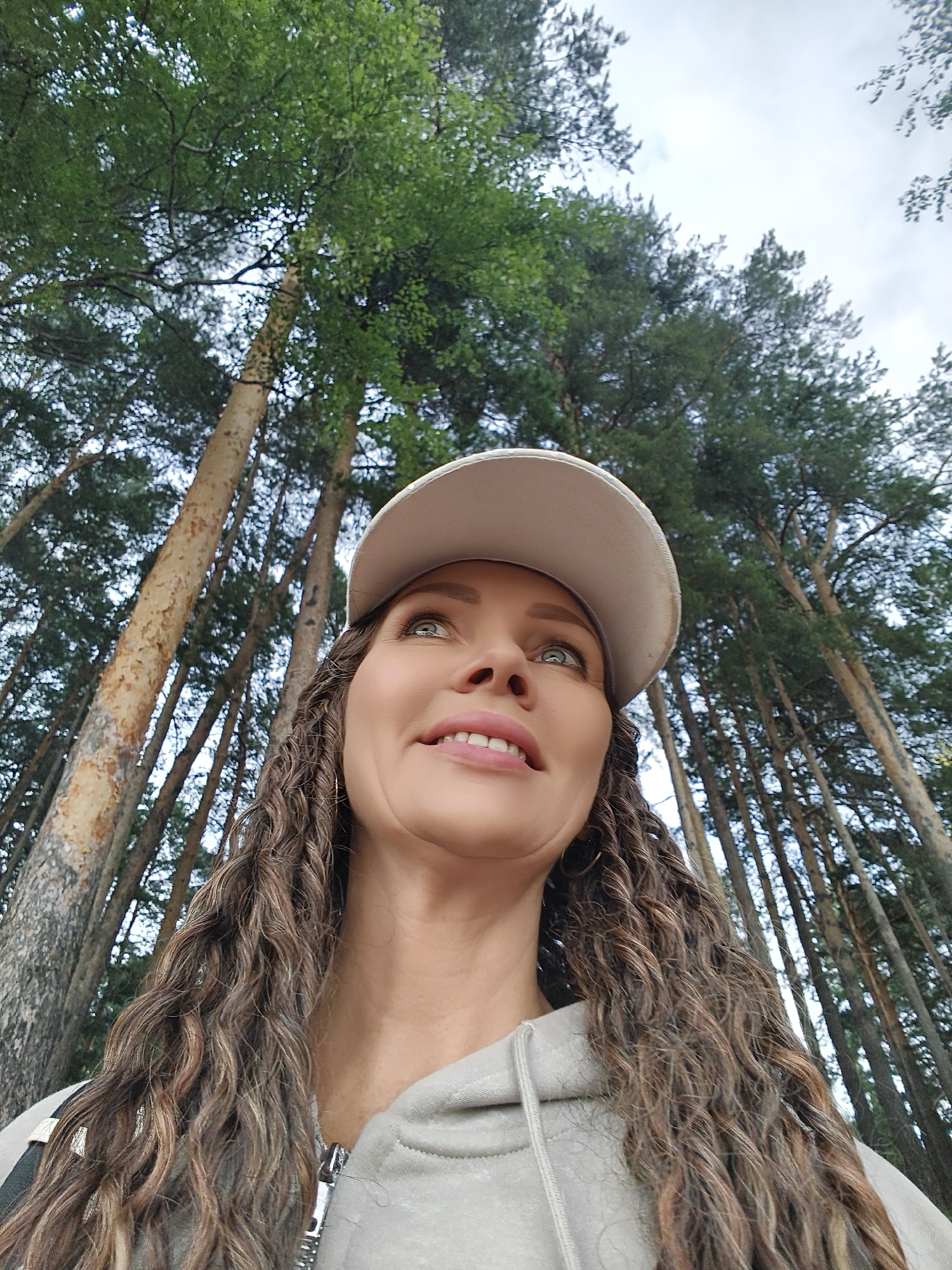 A low-angle, medium shot captures a woman looking upwards, with a bright smile and her eyes reflecting the light. She is wearing a light-colored baseball cap and a grey hooded jacket. Her long, curly brown hair cascades over her shoulders. The background features a dense forest of tall, slender trees, their trunks a mix of light and dark bark, with lush green foliage creating a canopy above. The sky is overcast with hints of blue peeking through. The scene suggests an outdoor activity, possibly a hike or a casual walk in the woods. The natural light and the woman's cheerful expression suggest it is daytime.