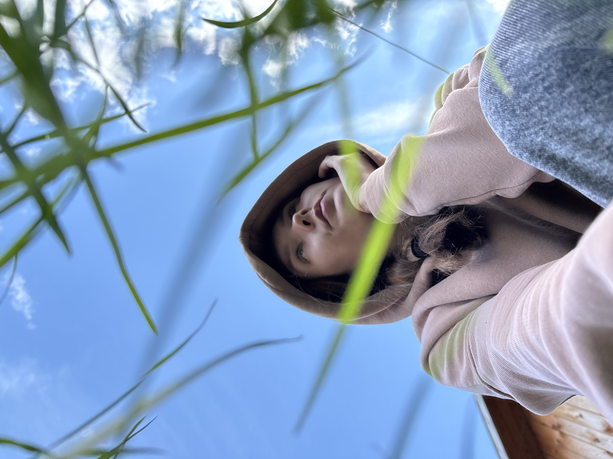A low-angle shot of a person wearing a beige hooded sweatshirt, with blue jeans visible on the upper right side of the frame. The person's face is partially obscured by the hood and the angle. Green blades of grass are visible in the foreground, slightly out of focus. The background is a light blue sky with scattered white clouds. A small wooden structure is visible at the bottom right. The location in the city of Italmas, Russia, is not discernible from the image.