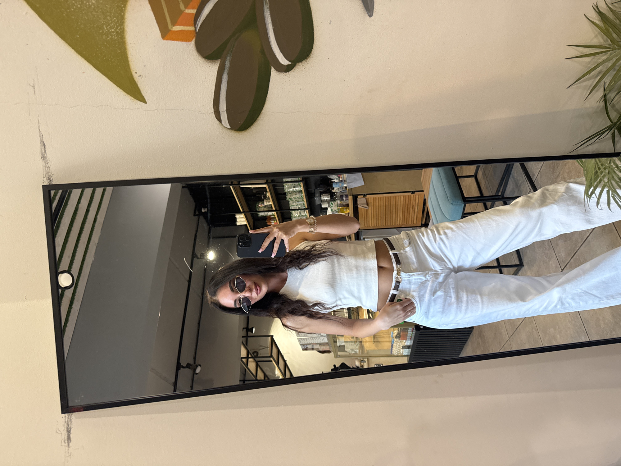 An indoor mirror selfie features a young woman posing in front of a full-length, black-framed mirror. The woman has long, dark, wavy hair and is wearing dark, cat-eye style sunglasses with gold-colored frames. She is dressed in a white ribbed crop tank top, white high-waisted wide-leg jeans, and a slim dark brown belt with a gold-toned rectangular buckle. A gold-colored watch or bracelet with multiple links is visible on her left wrist (right side of the image), and a ring is on her left-hand ring finger. Her left hand (right in the image) holds a dark-colored smartphone, possibly an iPhone with a triple camera array, positioned to capture the reflection. Her right hand (left in the image) is casually tucked into the front pocket of her jeans.
The reflection in the mirror shows a commercial interior space, potentially a cafe or retail establishment. Behind the woman, dark-framed open shelving units are visible, displaying various bottles, containers, and boxes. Below the shelving, a light-colored wooden slatted cabinet or counter is present. The ceiling reflection shows a white circular light fixture and recessed spot lighting. A tiled floor is also reflected. A blue upholstered stool with a black metal frame is reflected near the bottom right of the mirror.
The wall outside the mirror frame is a light cream or beige color. To the right of the mirror, a large, abstract mural depicts stylized brown, oval shapes with white stripes, an olive-green leaf-like element, and an orange geometric shape. Multiple fine, vertical hairline cracks are present on this wall, particularly around the mural. Near the top left corner of the mirror, faint gray smudges are visible on the wall. A small, leafy green plant is partially visible on the floor in the bottom right corner of the scene, outside the mirror.