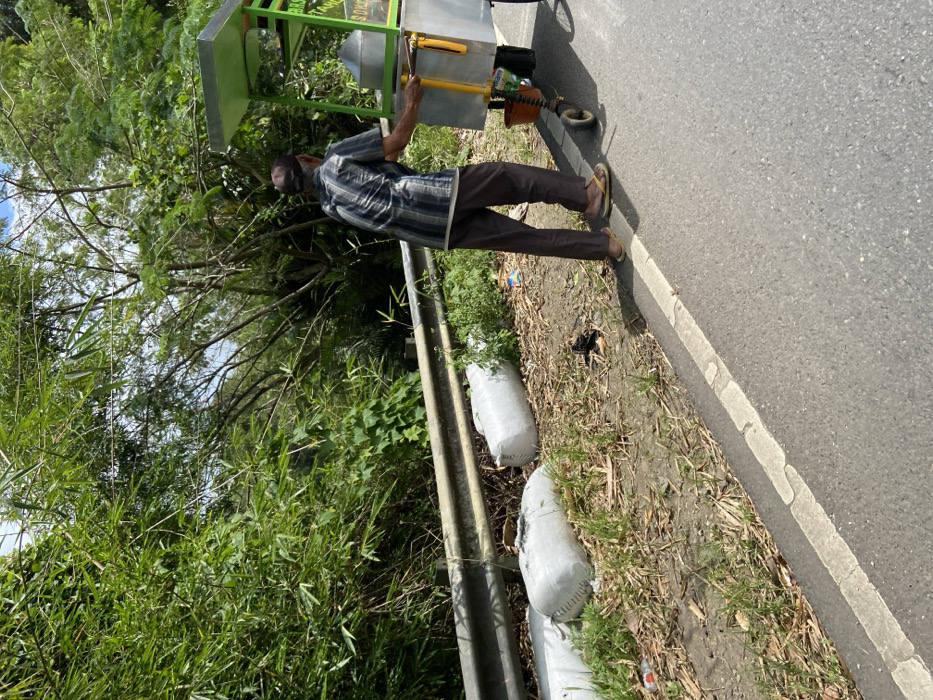A man in a striped shirt and brown pants is standing beside a green and white food cart on the side of a paved road in Cipatat, Indonesia. The road has a white dividing line. The man is looking down at the cart. The cart is on the side of the road, next to a metal guardrail. There are three white bags on the ground near the guardrail. There are green bushes and trees behind the man and the cart. The location is approximately -6.787, 107.434. The man is likely a vendor selling food from the cart.