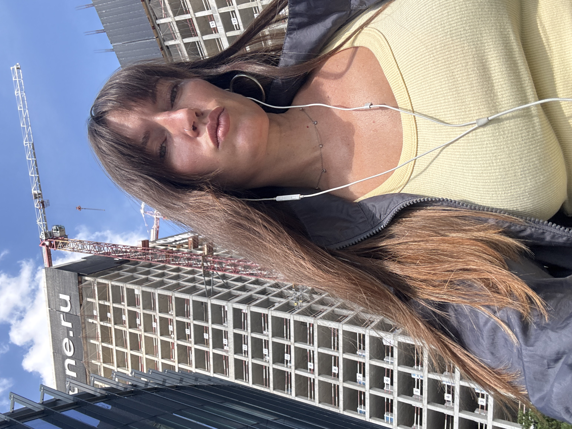 In Khimki, Russia, a woman with long, brown hair and bangs is captured in a selfie against a backdrop of urban construction. She is wearing a light yellow tank top and a dark jacket. White earbuds are visible, with one earpiece resting in her ear and the cord trailing down. A delicate necklace with small, sparkling charms adorns her neck. She has a silver hoop earring in her left ear and a small nose ring. The background features a partially constructed, multi-story building with visible concrete framework and numbered floors, indicating ongoing development. A large red and white construction crane is prominently positioned to the right, reaching towards the blue sky dotted with white clouds. The overall scene conveys a sense of an active construction site within a city environment.